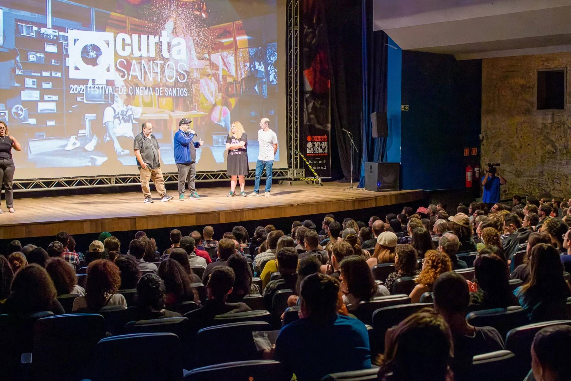 Pessoas no palco de um auditório durante o Festival Curta Santos 2021, com a plateia lotada e telão exibindo logo do evento.