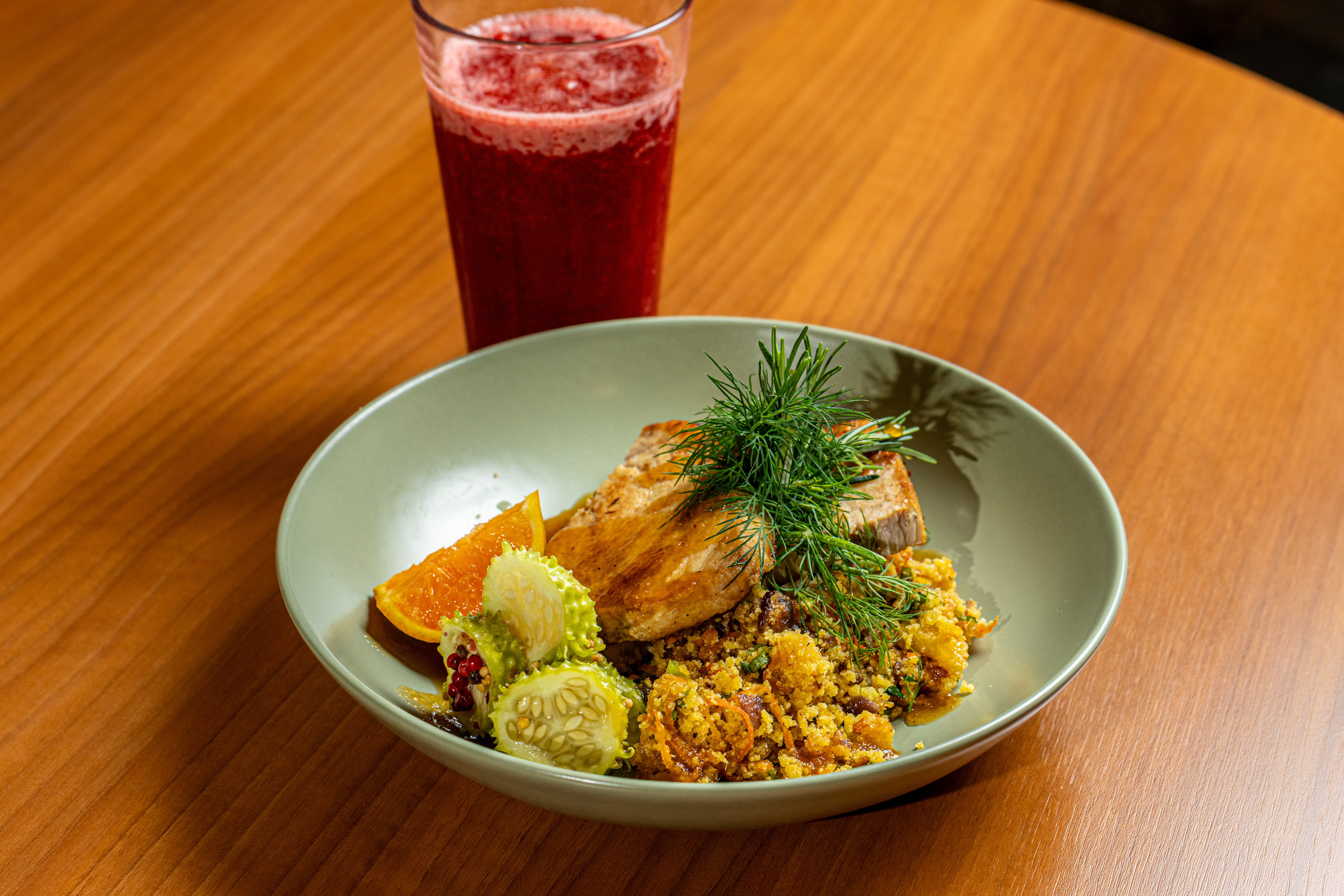 Filé grelhado com farofa, fatias de laranja e kiwano, decorado com endro, acompanhado de suco vermelho em copo alto sobre mesa de madeira.