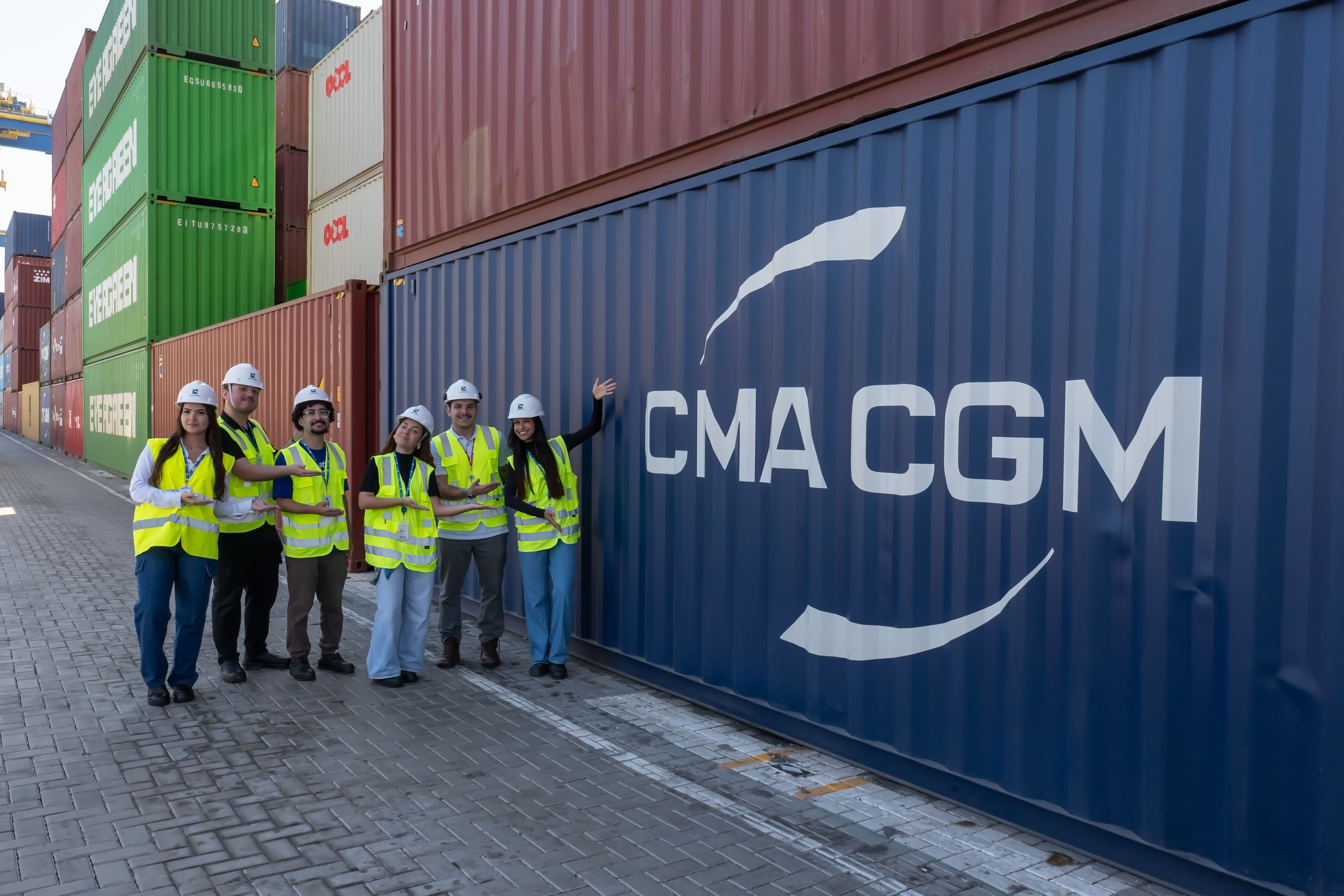 Profissionais com capacetes e coletes de segurança sorrindo e apontando para contêiner CMA CGM azul em terminal de cargas.