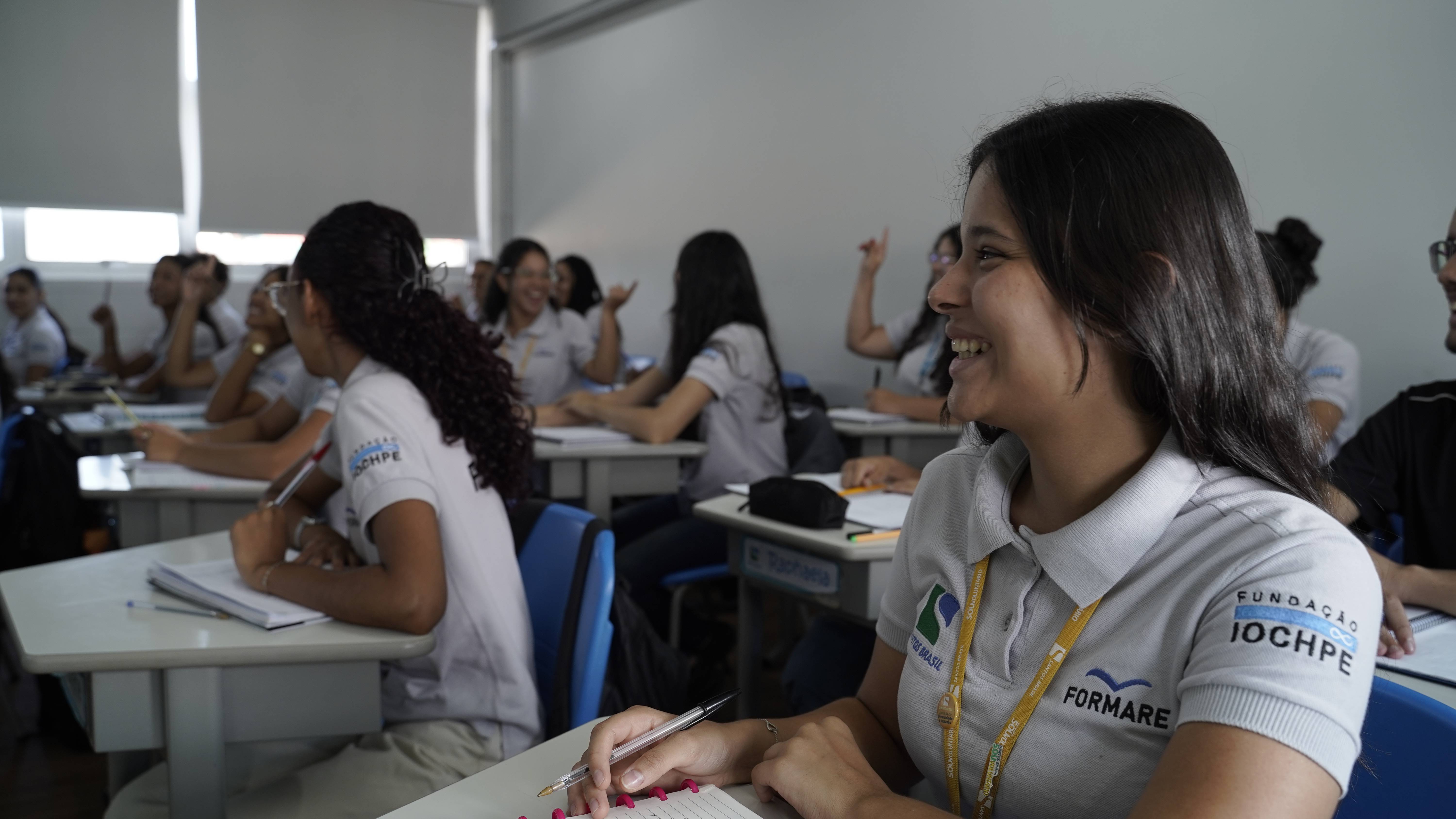 Jovens em sala de aula, uma estudante sorrindo em primeiro plano, usando camisa com logos da Fundação IOCHPE e Formare.