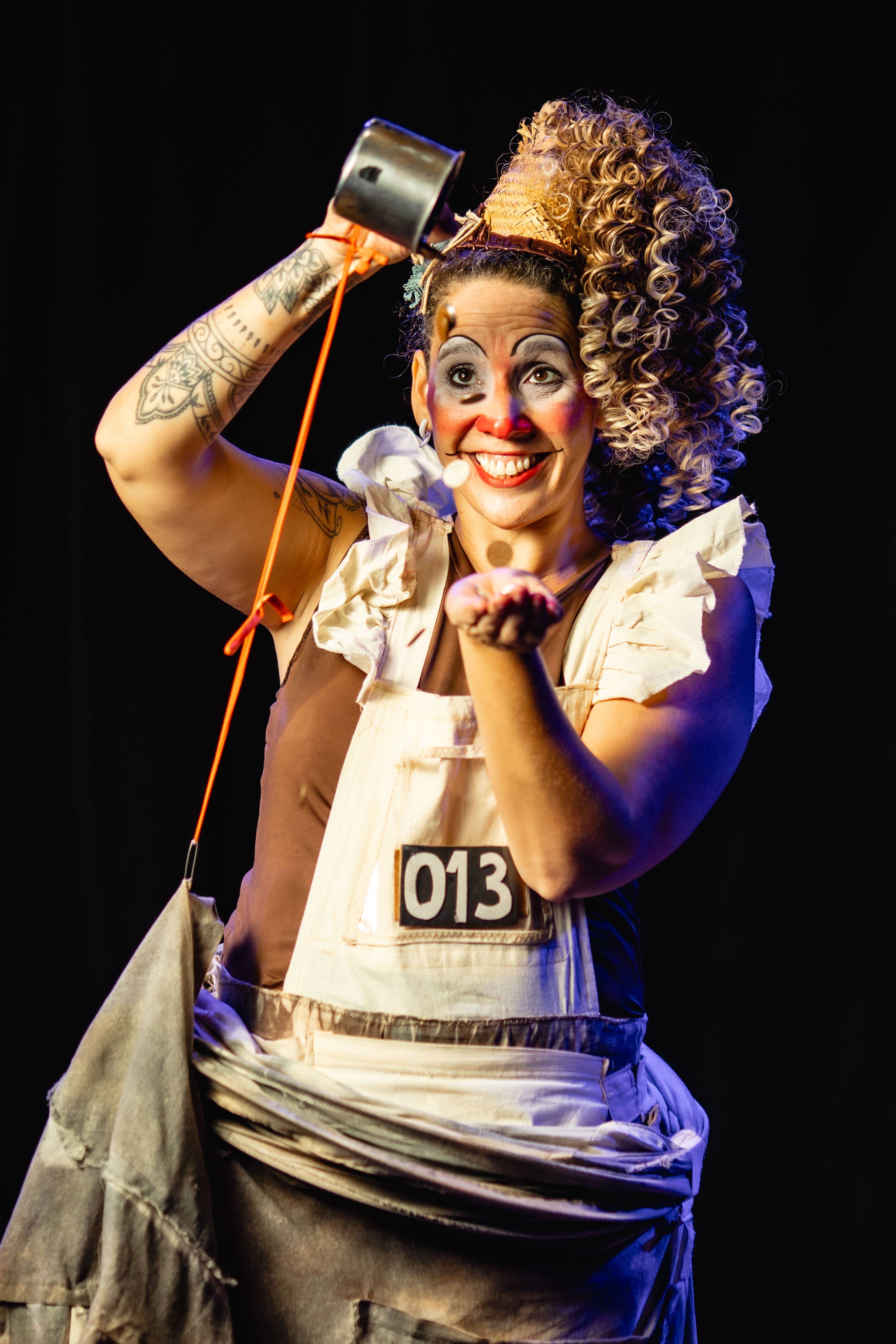 Palhaça sorridente com maquiagem teatral e cabelo encaracolado, derramando confetes de uma caneca enquanto estende a mão.