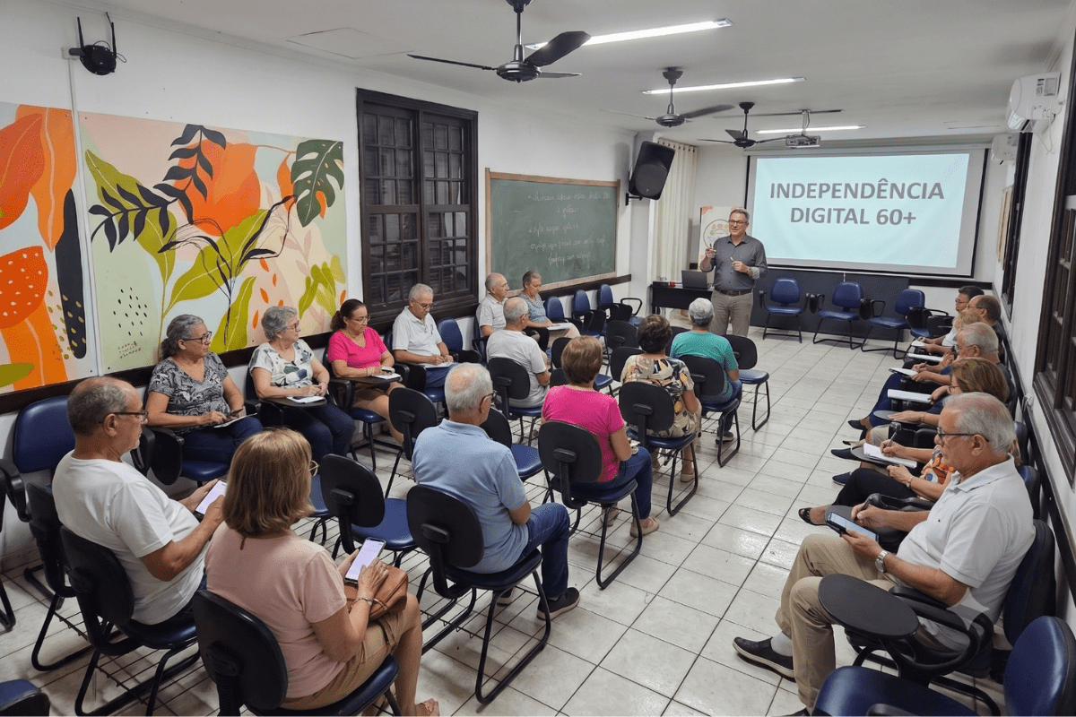 Grupo de idosos participa de palestra sobre "Independência Digital 60+", com instrutor gesticulando em frente à tela.