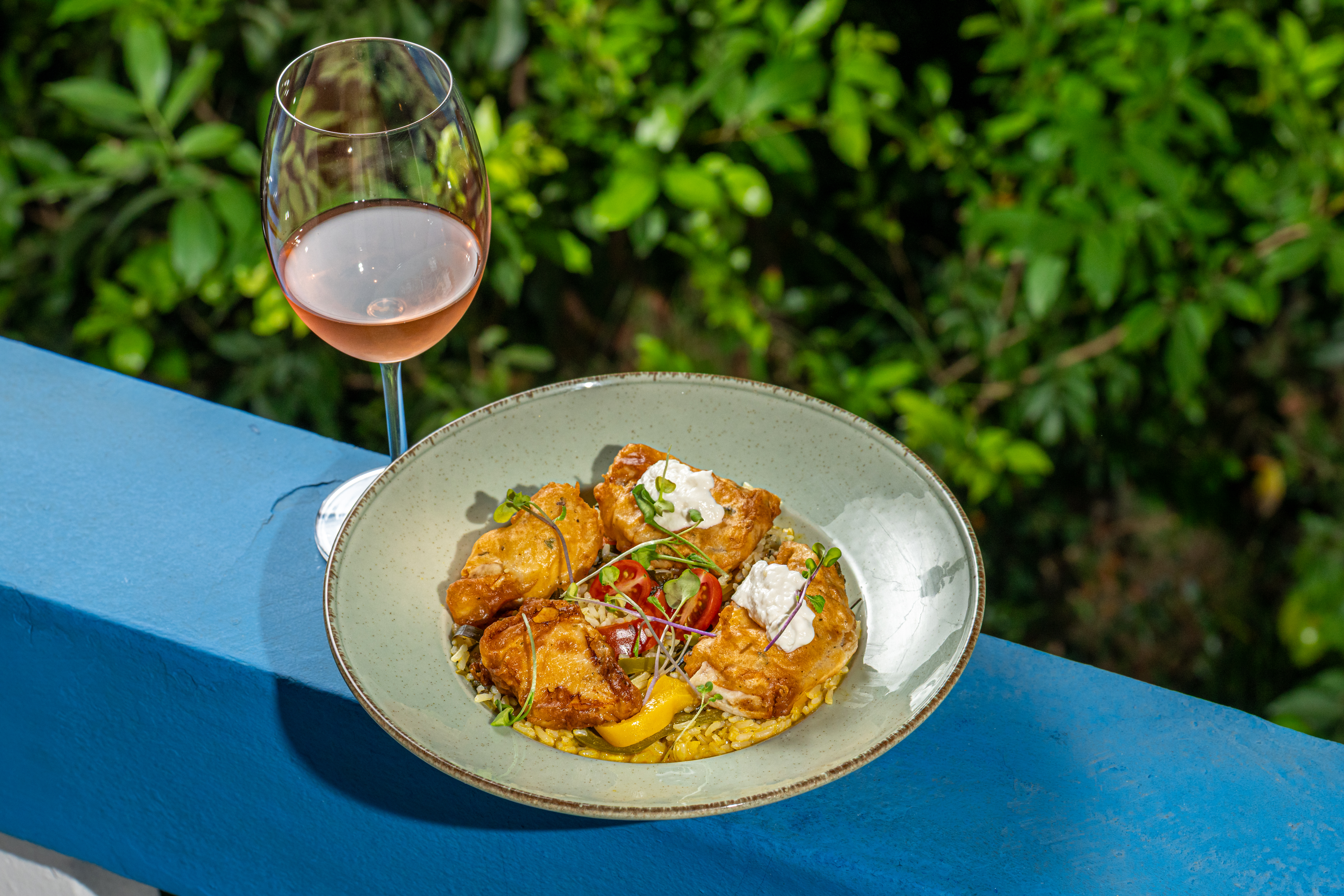 Prato com peixe frito empanado sobre arroz, guarnecido com tomate, pimentão e microgreens, acompanhado de taça de vinho rosé.