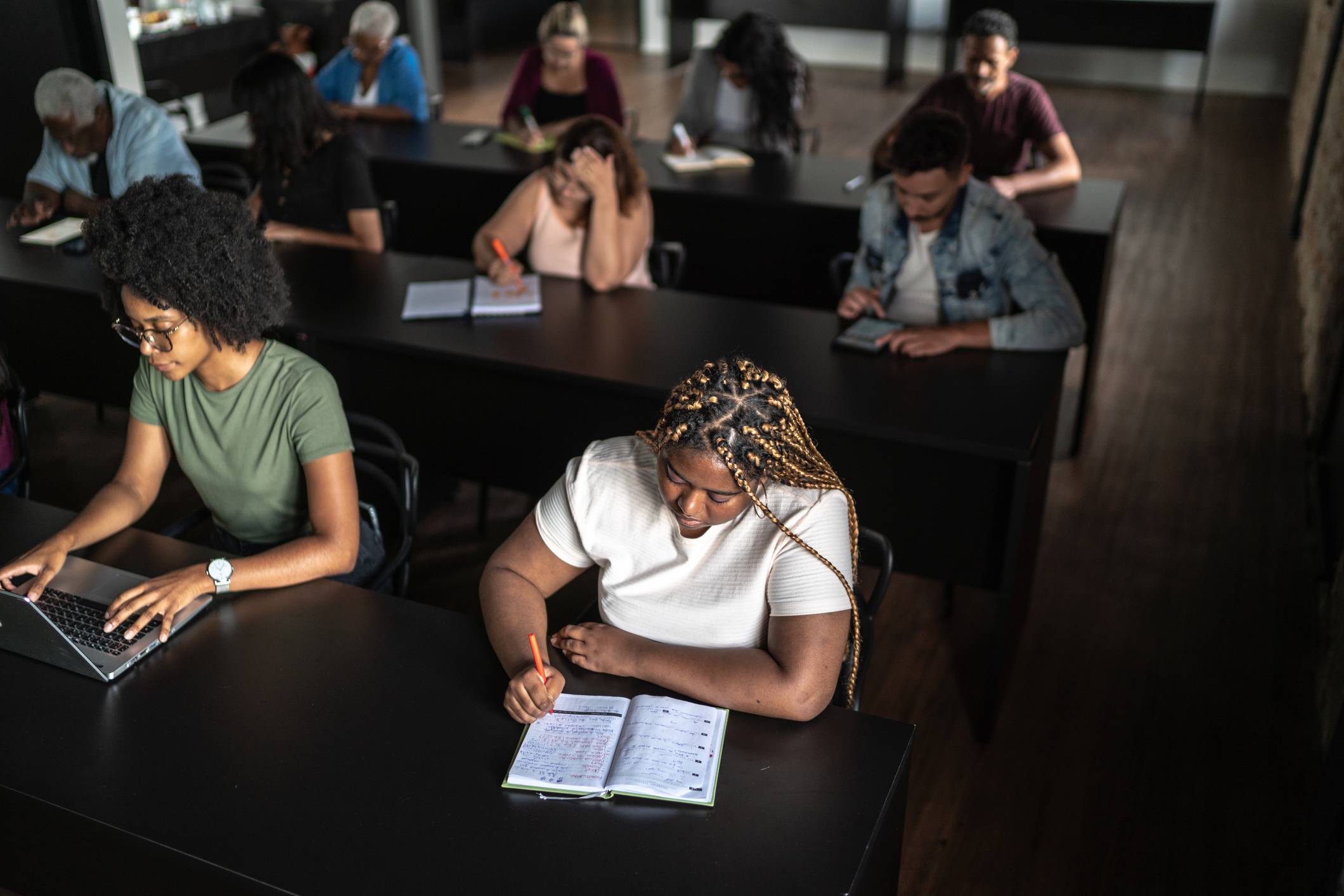Alunos adultos diversos, incluindo uma mulher no laptop e outra escrevendo, aprendem em sala de aula com mesas individuais.