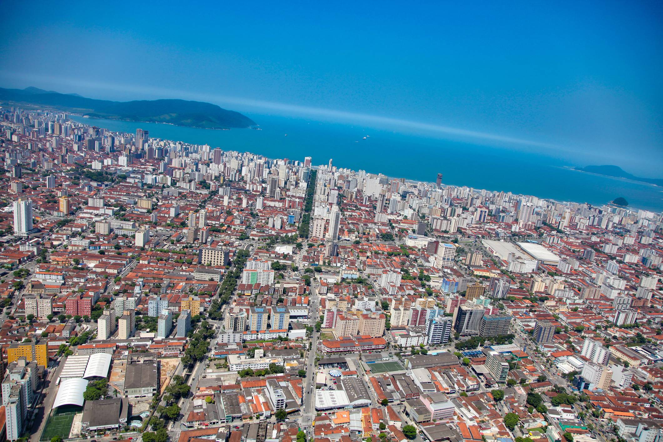 Vista aérea panorâmica de uma cidade costeira densamente construída com arranha-céus, oceano azul e montanhas distantes.