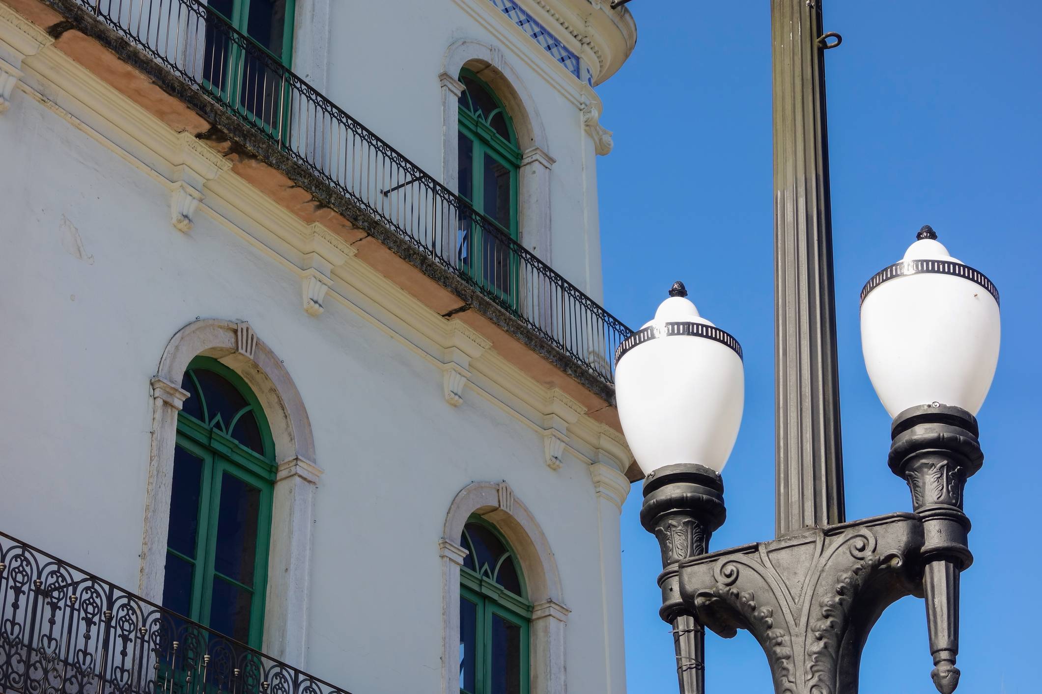 Fachada branca de edifício histórico com janelas verdes arqueadas e varandas de ferro forjado, ao lado de poste de luz ornamentado contra céu azul.