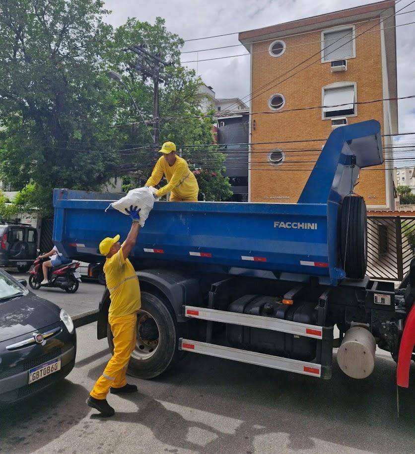 Dois trabalhadores de uniforme amarelo carregam saco de resíduos em caminhão basculante azul Facchini, em rua urbana.