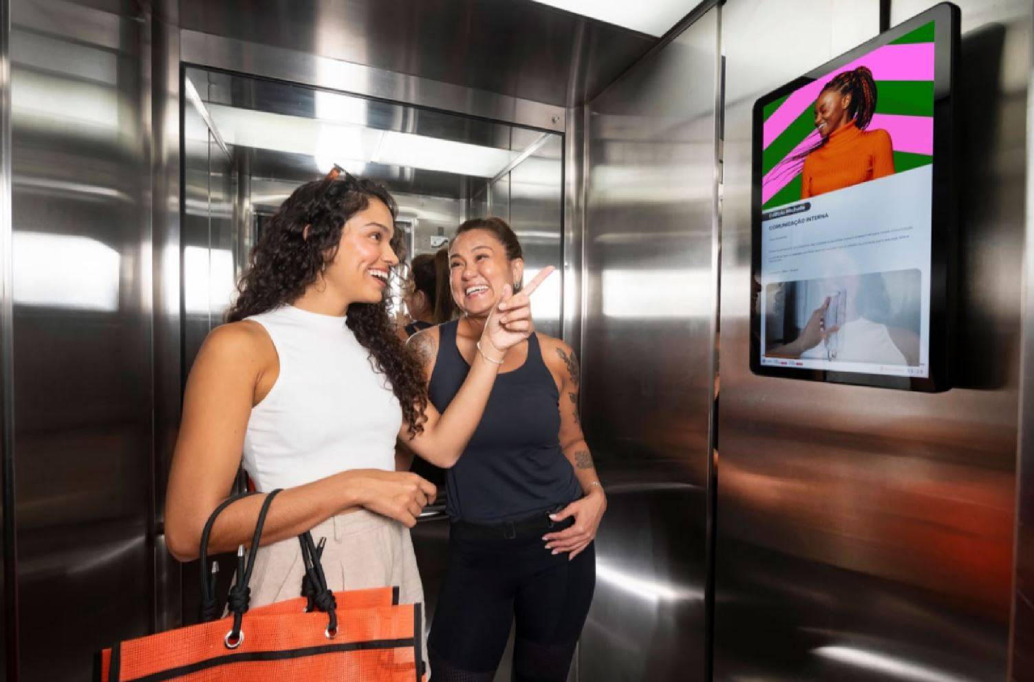 Duas mulheres rindo e interagindo dentro de um elevador moderno, uma apontando para uma tela digital exibindo comunicação interna.