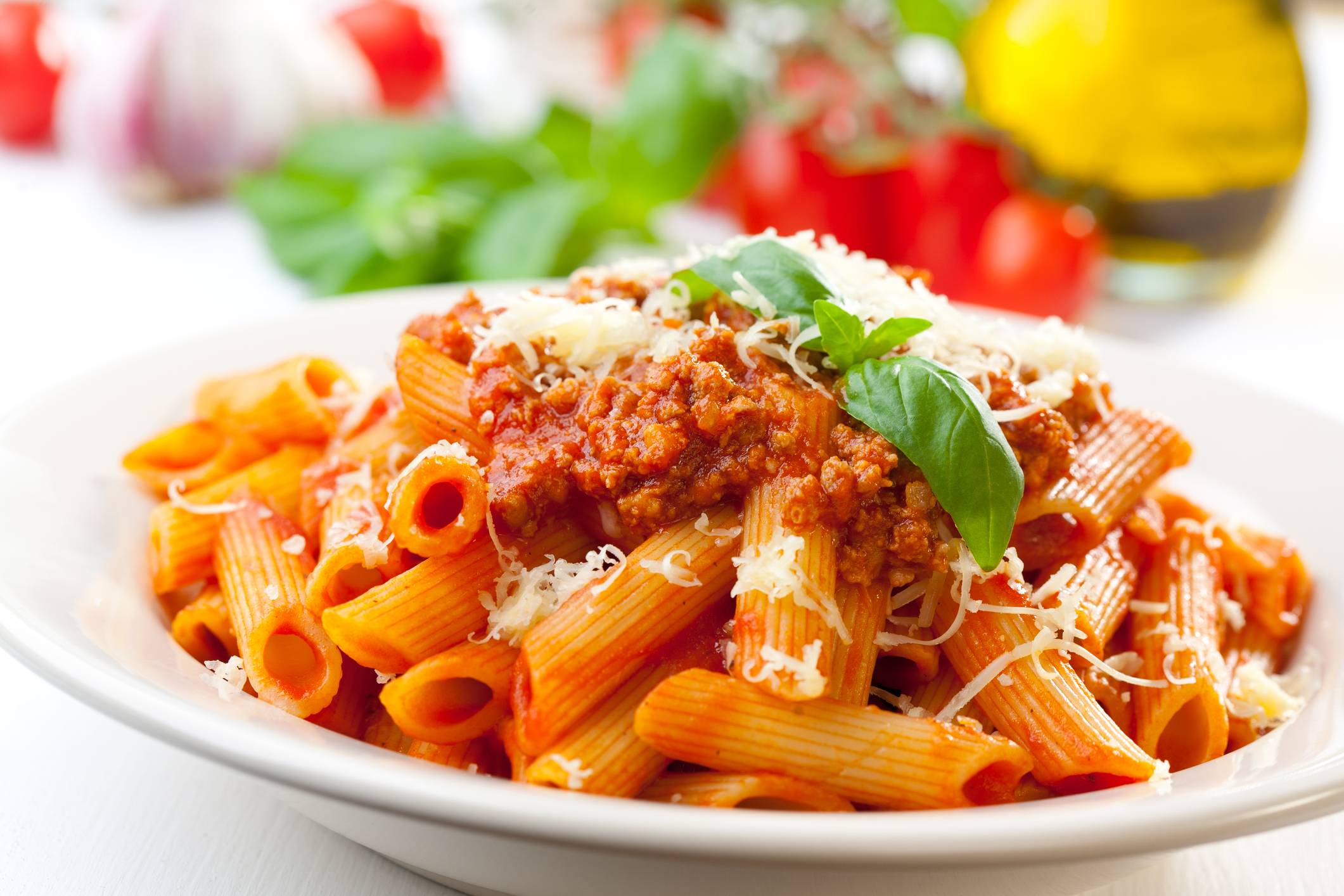 Penne pasta com molho bolonhesa, queijo ralado e manjericão fresco em prato branco, com ingredientes ao fundo.