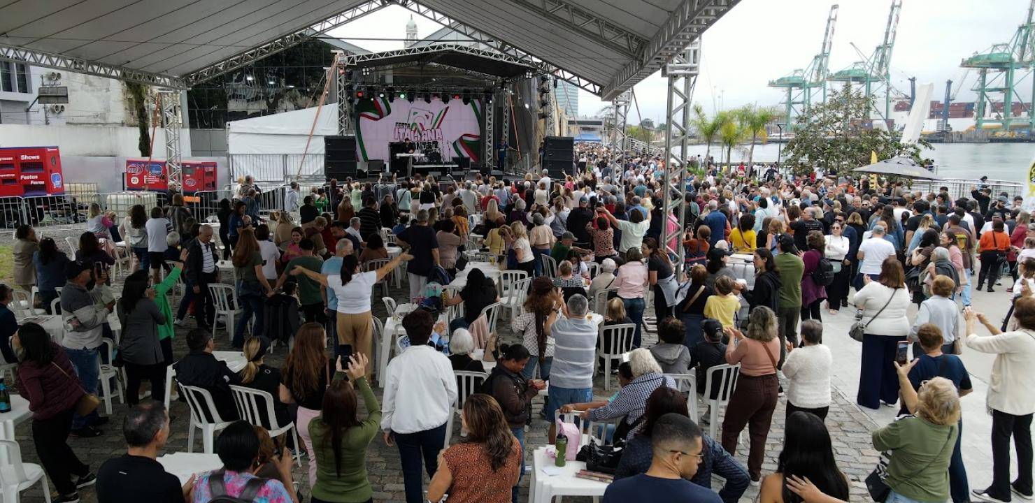 Multidão em Festa Italiana com palco, música ao vivo e guindastes portuários ao fundo durante o evento.