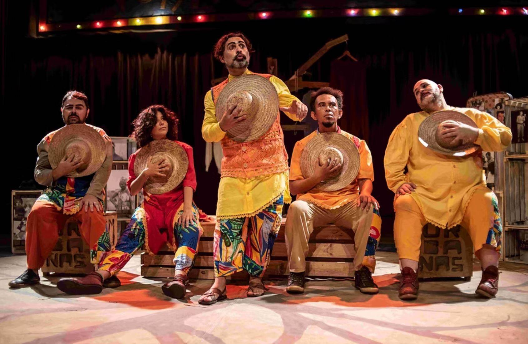 Cinco artistas de teatro em figurinos coloridos segurando chapéus de palha no palco, com luzes ao fundo.