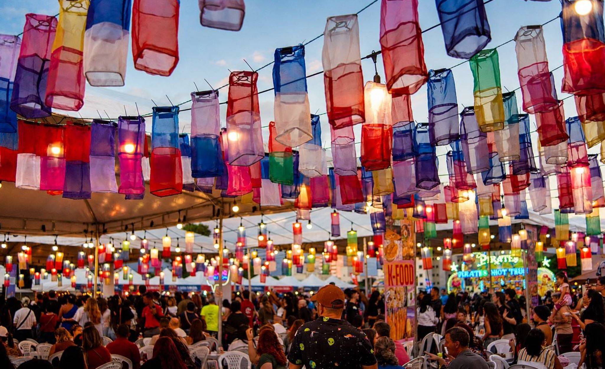 Festival noturno vibrante com centenas de lanternas coloridas translúcidas penduradas sobre uma multidão em barracas de comida.