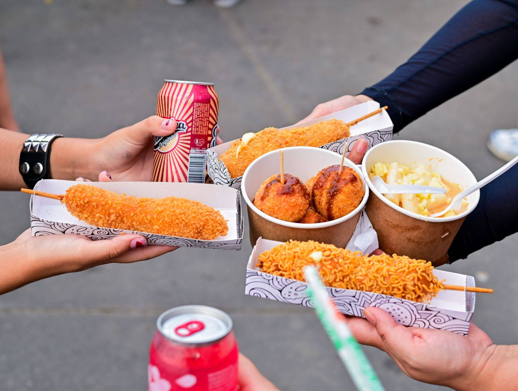 Pessoas segurando variedade de street food coreana: corndogs fritos, corndog de ramen, bolinhos e salada de macarrão, com refrigerantes.