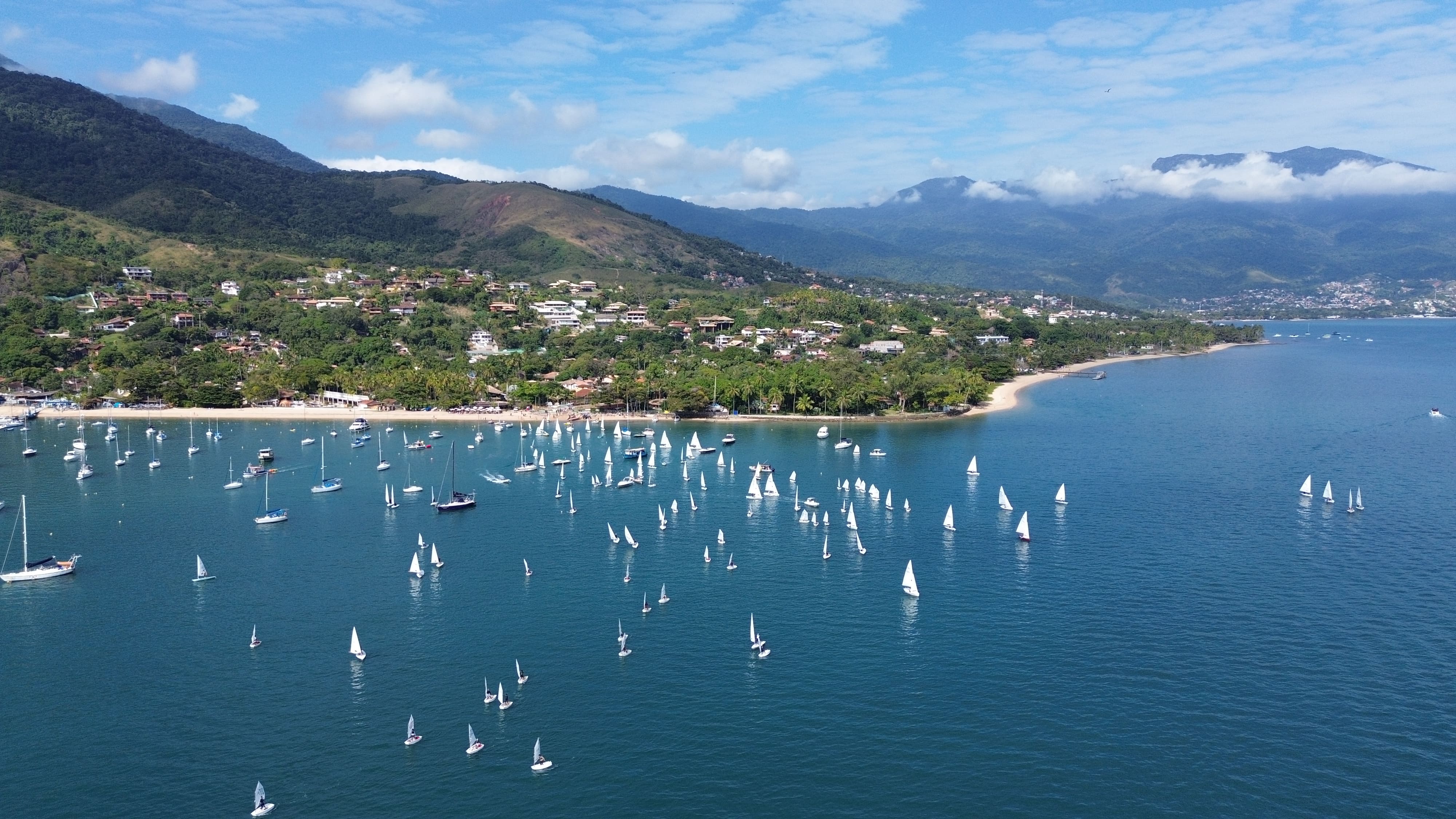 Vista aérea de uma baía com dezenas de veleiros em águas azuis, casas na encosta verdejante e montanhas ao fundo.