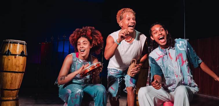 Três jovens sorrindo e cantando, um tocando pandeiro e outro um caxixi, em um palco escuro com conga ao lado.