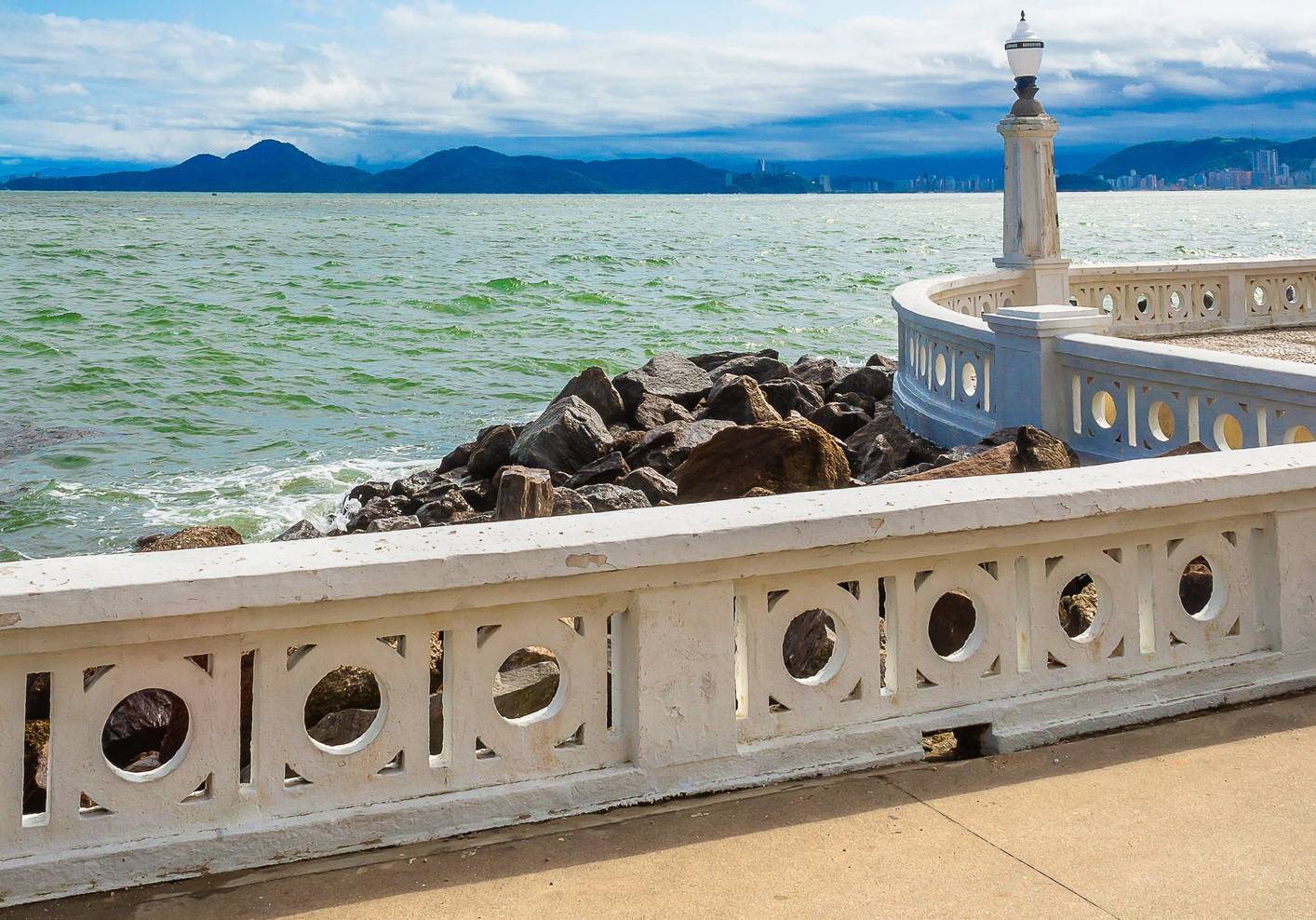 Passeio à beira-mar com balustrada branca, lampião, rochas no mar verde-azulado e montanhas com cidade ao fundo