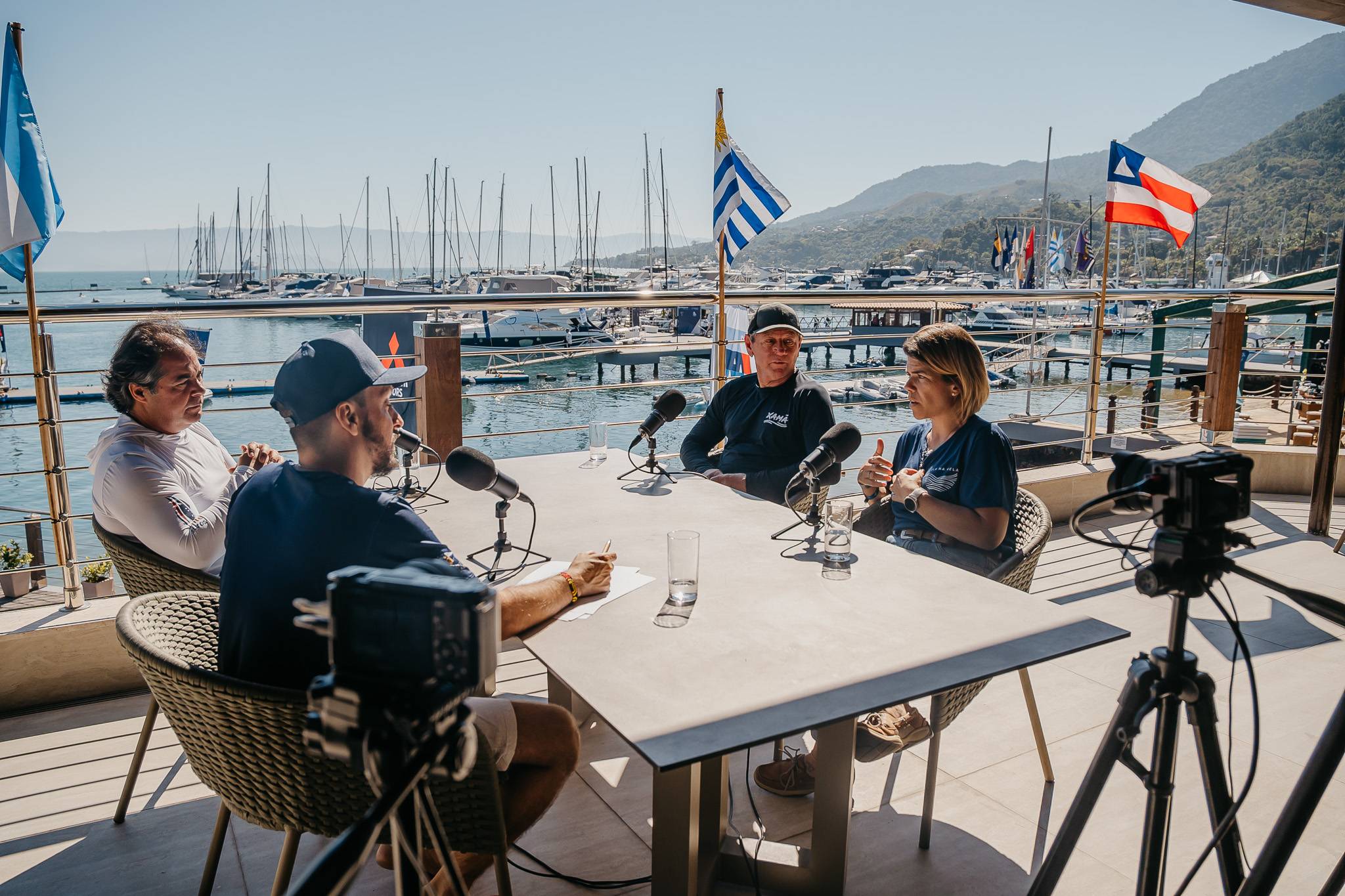 Quatro pessoas gravando podcast em varanda. Vista para marina com iates, montanhas. Microfones, câmera, bandeiras Argentina, Uruguai, Bahia.