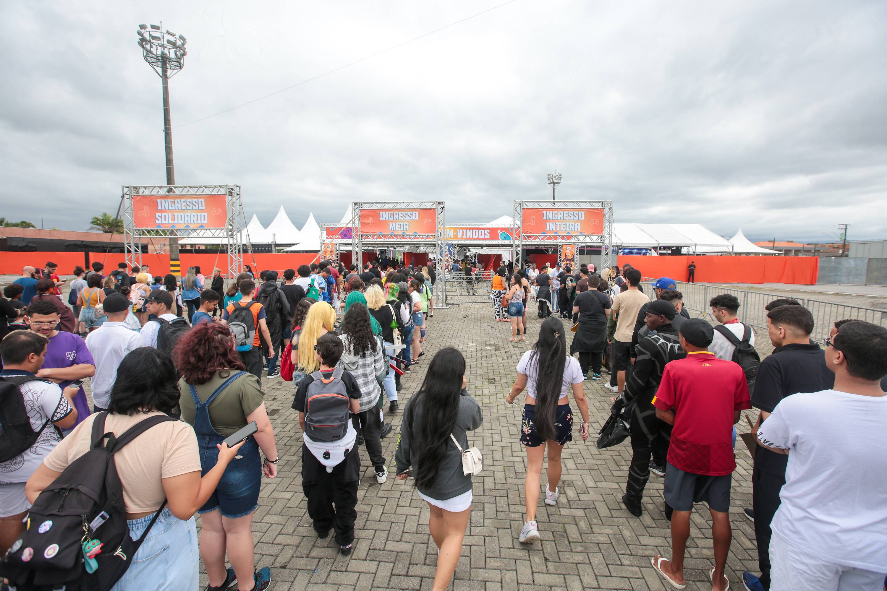 Pessoas em uma entrada de evento com placas indicando diferentes tipos de ingresso: solidário, meia e inteira. Multidão aguardando em filas.