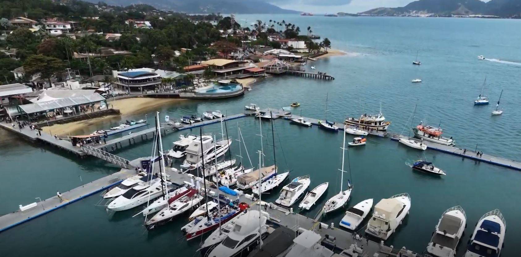 Marina movimentada com iates e barcos atracados, píeres cheios de pessoas, edifícios à beira-mar e montanhas ao fundo.