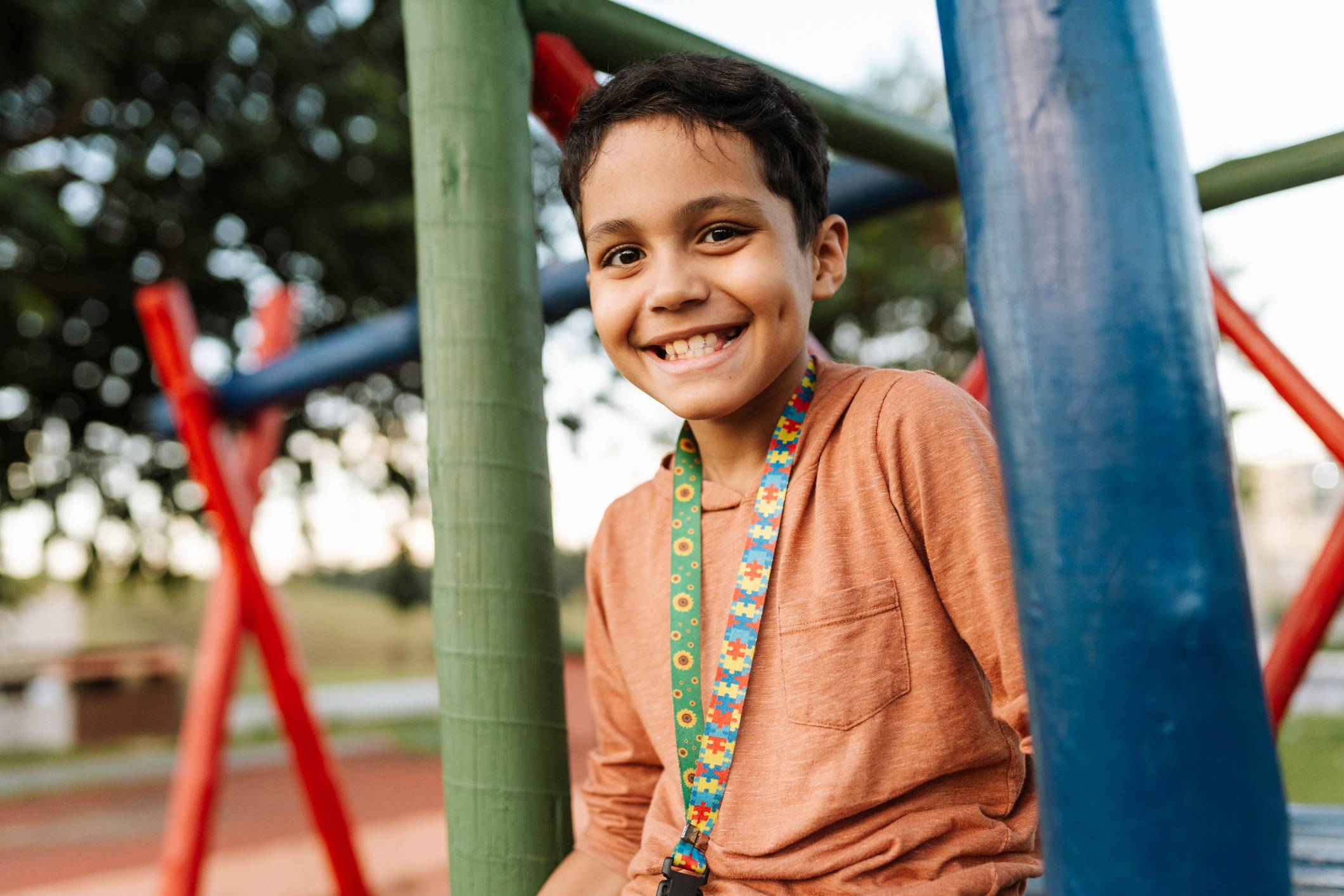 Menino feliz sorridente com dimples, camisa laranja e cordão de quebra-cabeças e girassóis em parquinho.