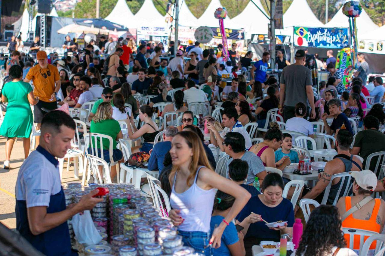 Muitas pessoas comendo e socializando em mesas brancas sob tendas em um evento diurno ao ar livre movimentado.