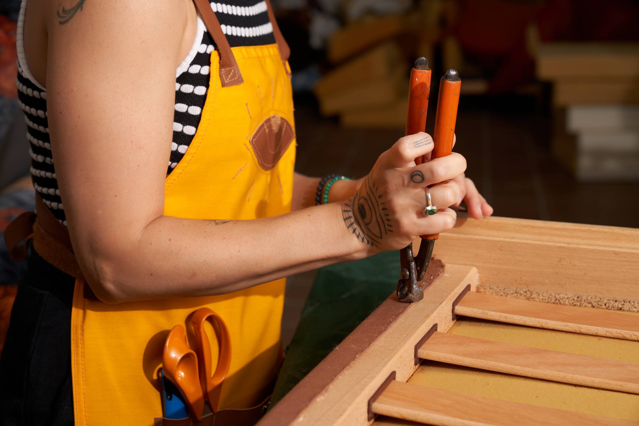 Mãos de artesã com avental amarelo e tatuagens, usando alicate para fixar material em estrutura de madeira. Foco no trabalho manual.