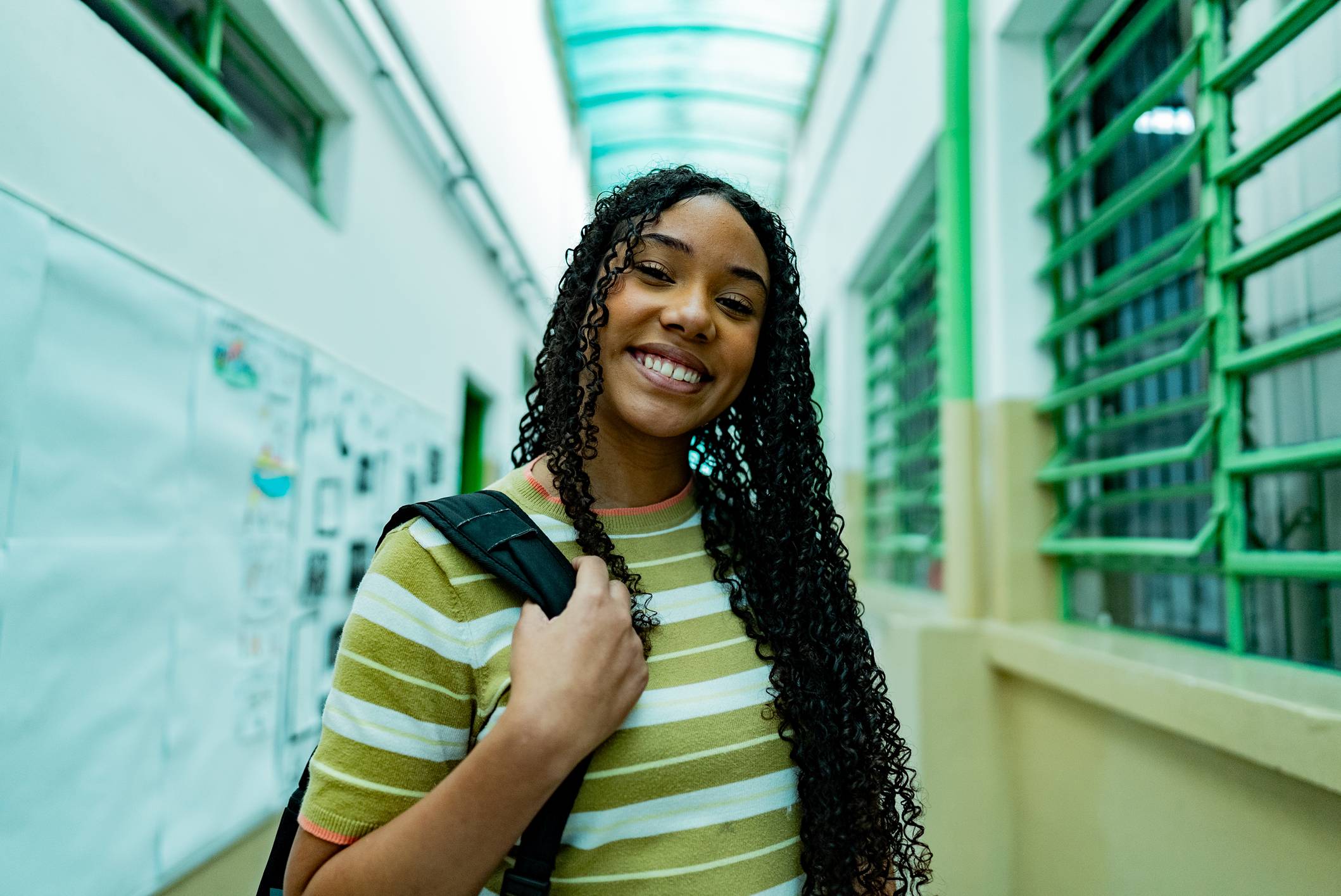 Jovem mulher negra de cabelos encaracolados sorrindo na câmera, com mochila, em corredor iluminado com janelas e quadro de avisos.