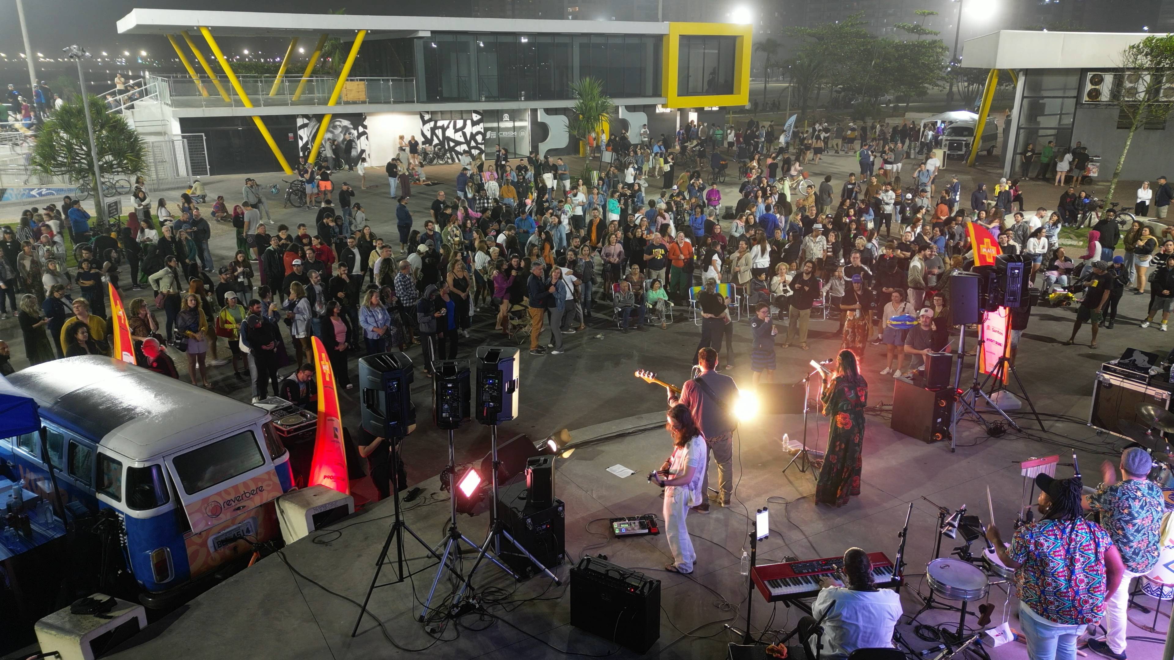 Banda se apresentando ao vivo para uma multidão em um evento noturno, com Kombi vintage e arquitetura moderna ao fundo.