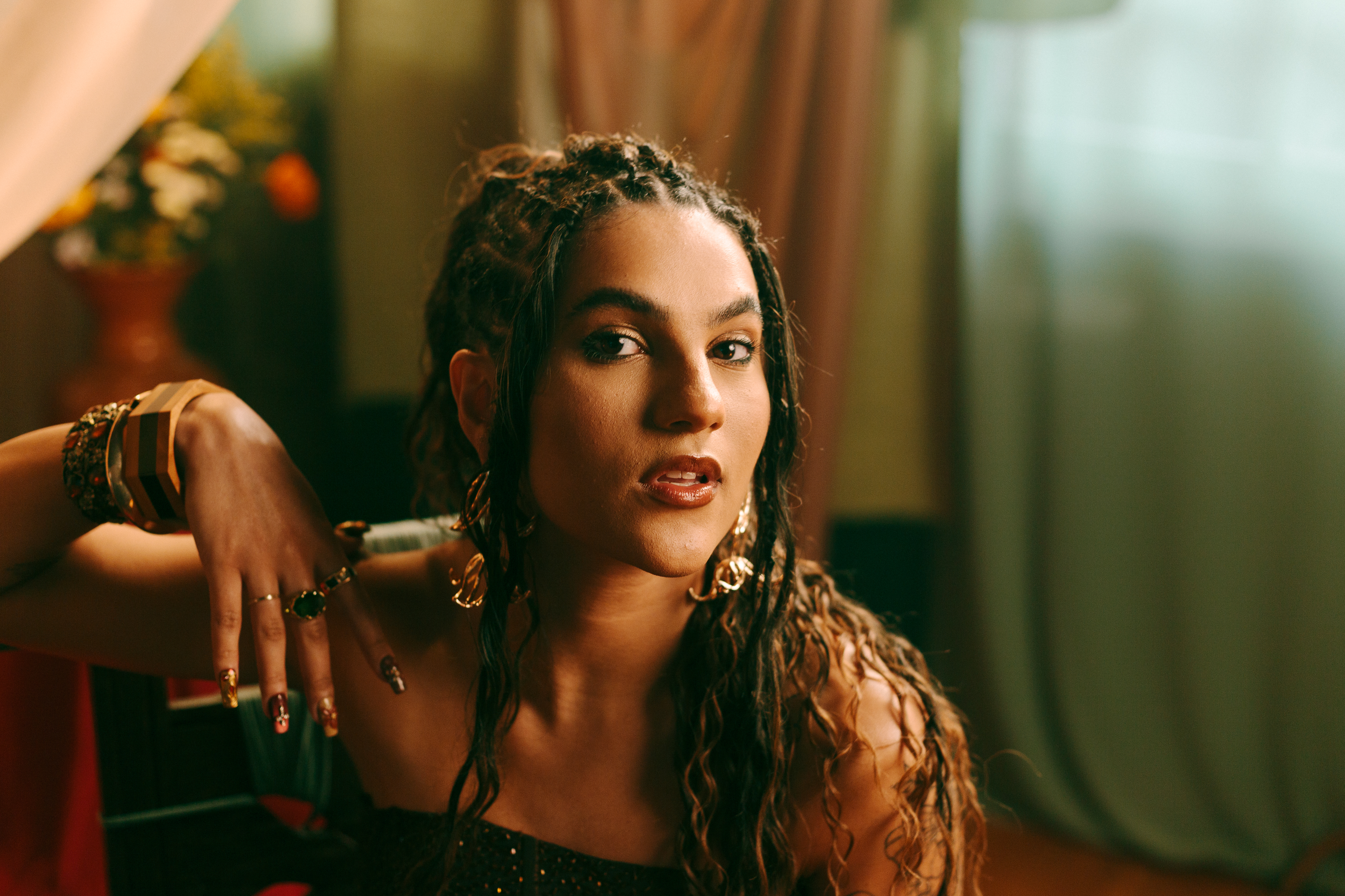 Mulher com tranças, maquiagem destacada nos olhos e lábios, brincos grandes e pulseiras de ouro, olhando fixamente para a câmera.