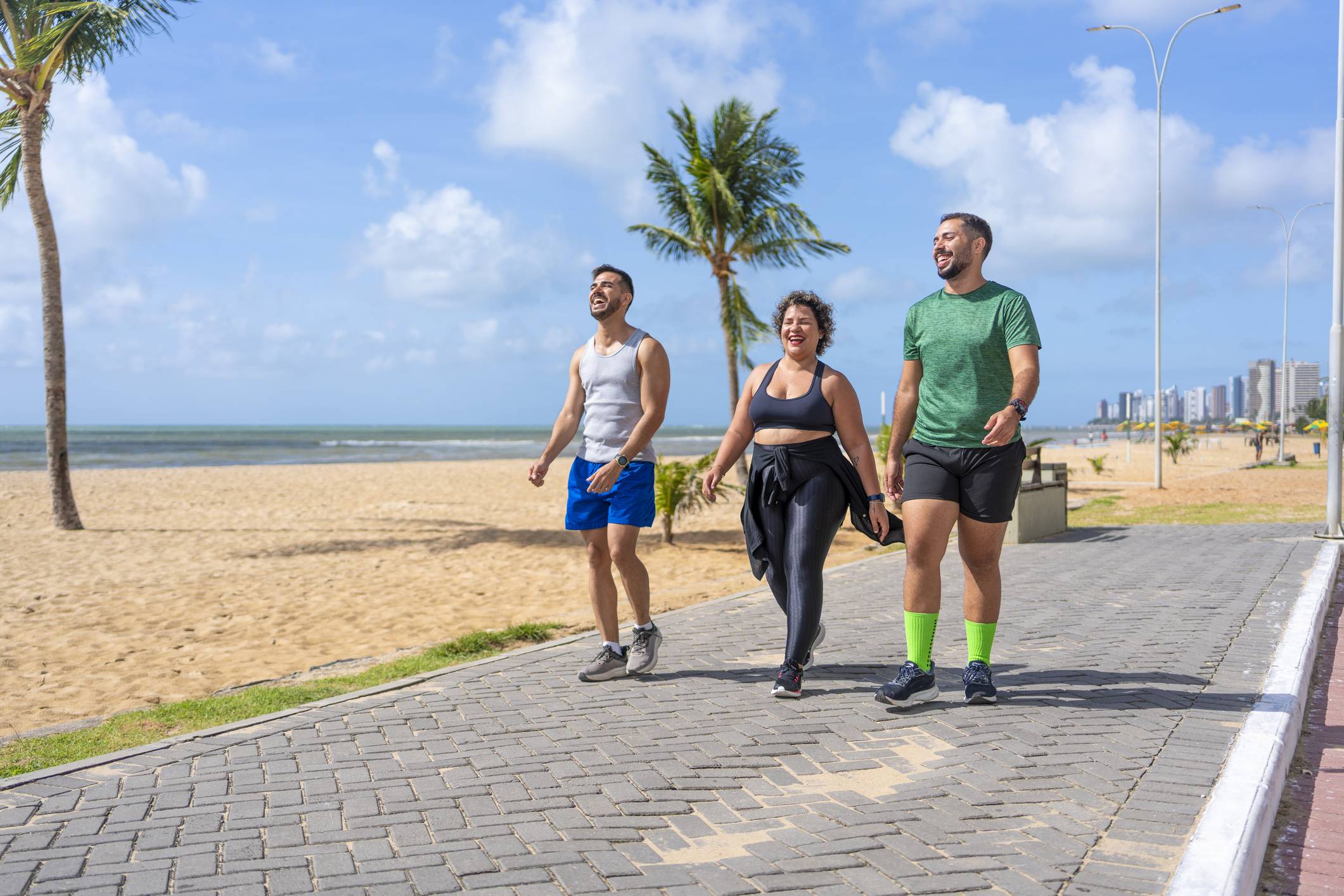 Três pessoas (dois homens, uma mulher) caminhando em calçadão à beira-mar com palmeiras, praia e oceano ao fundo, sob céu azul.