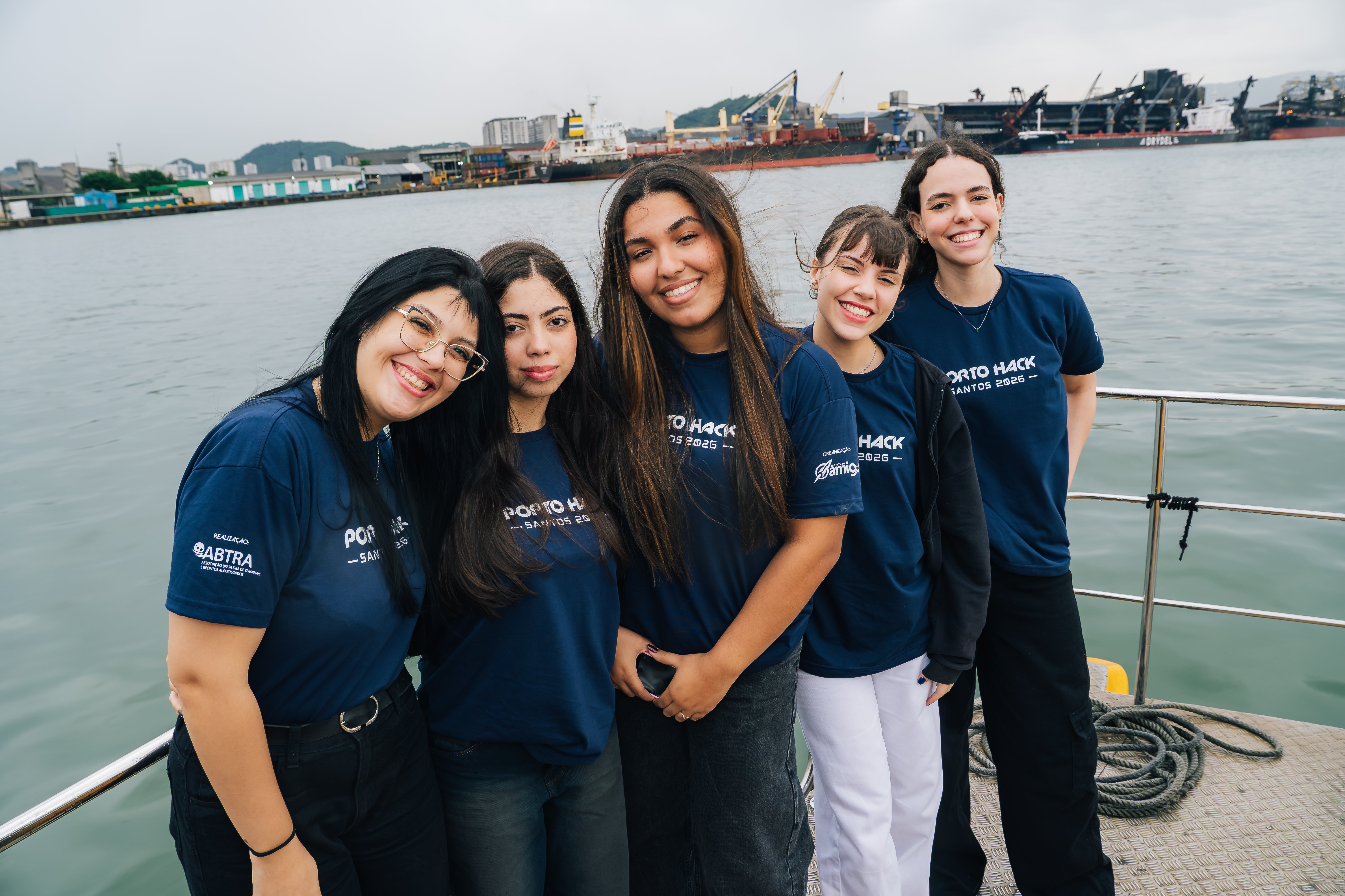 Cinco jovens sorrindo, usando camisetas "Porto Hack Santos 2026" em um barco, com porto e navios ao fundo.