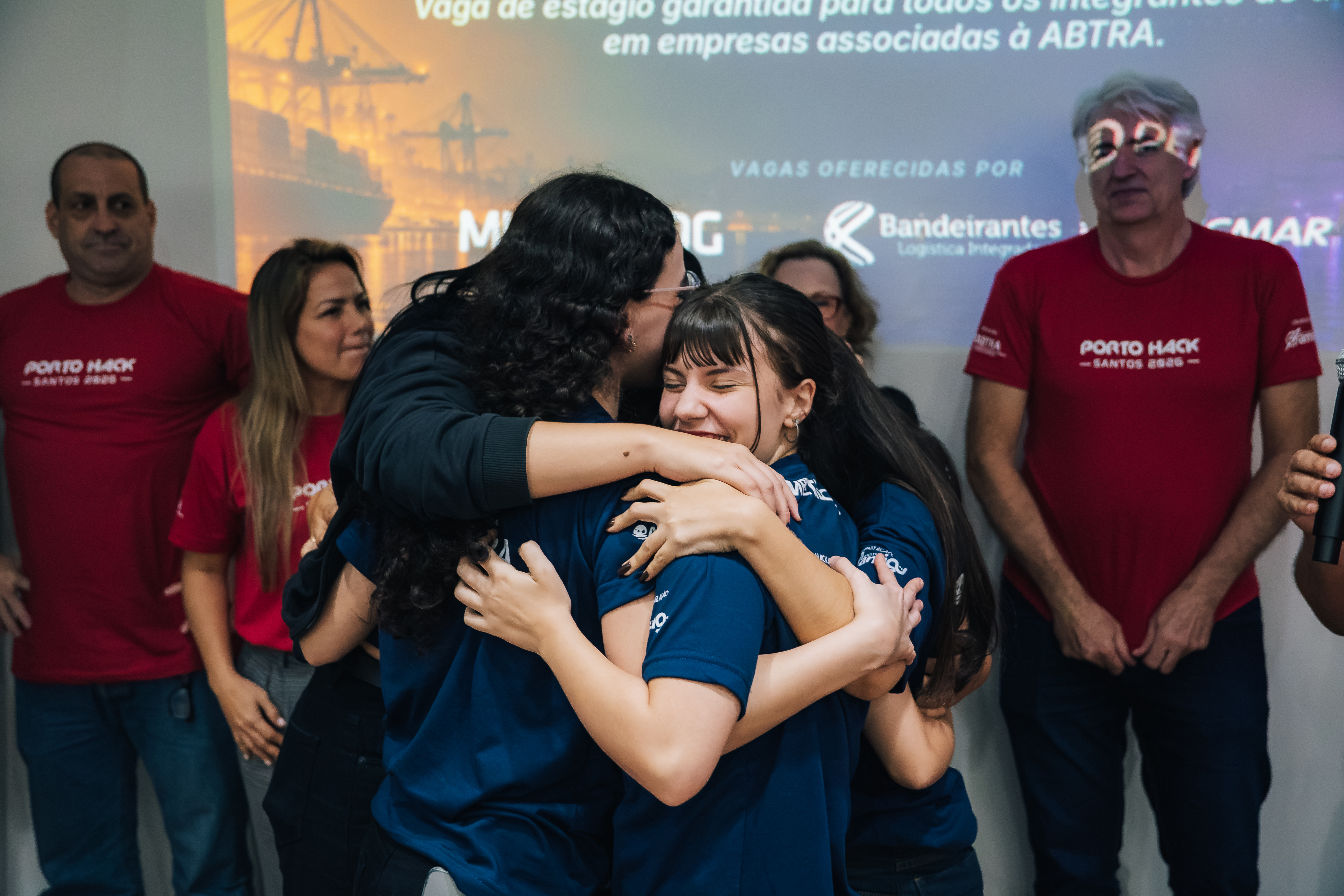 Duas jovens se abraçam com emoção em evento, enquanto outras pessoas as observam. Ao fundo, uma projeção sobre oportunidades de estágio.