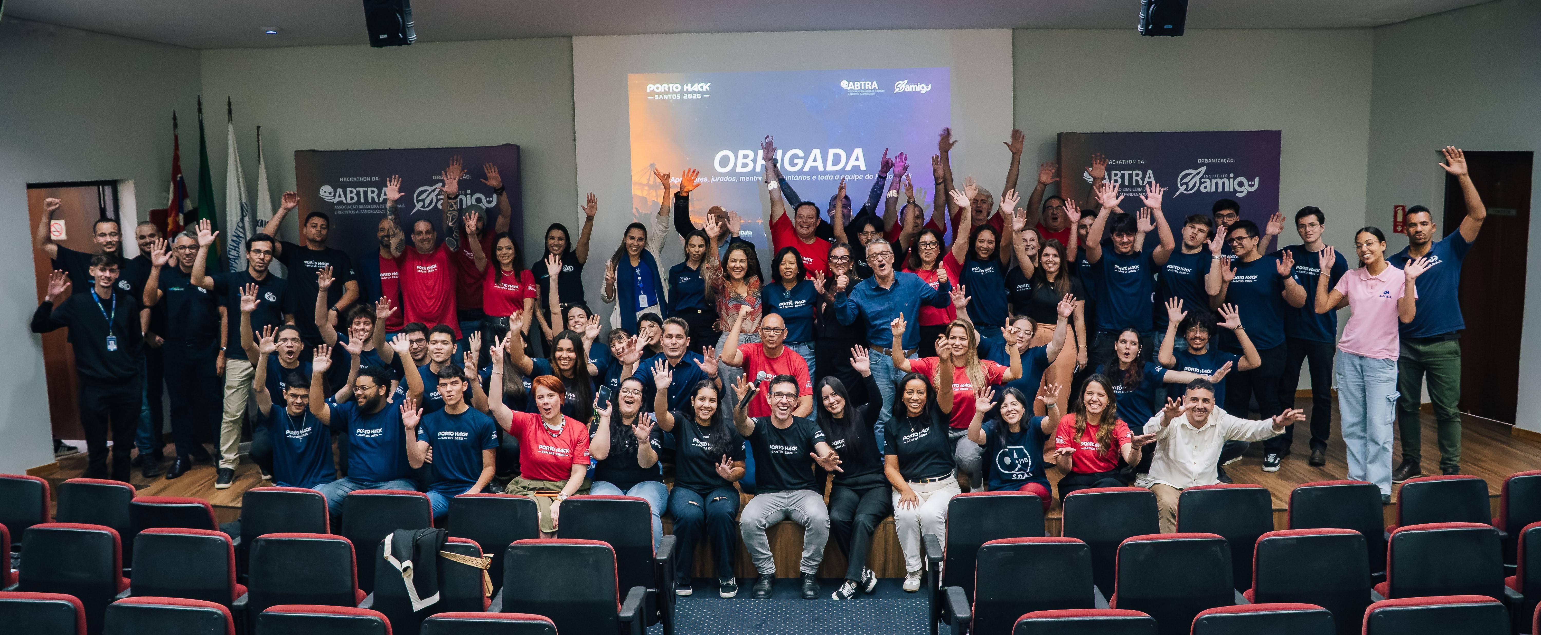 Grupo de participantes e organizadores do Porto Hack Santos 2026 em pose animada, acenando e sorrindo em auditório.