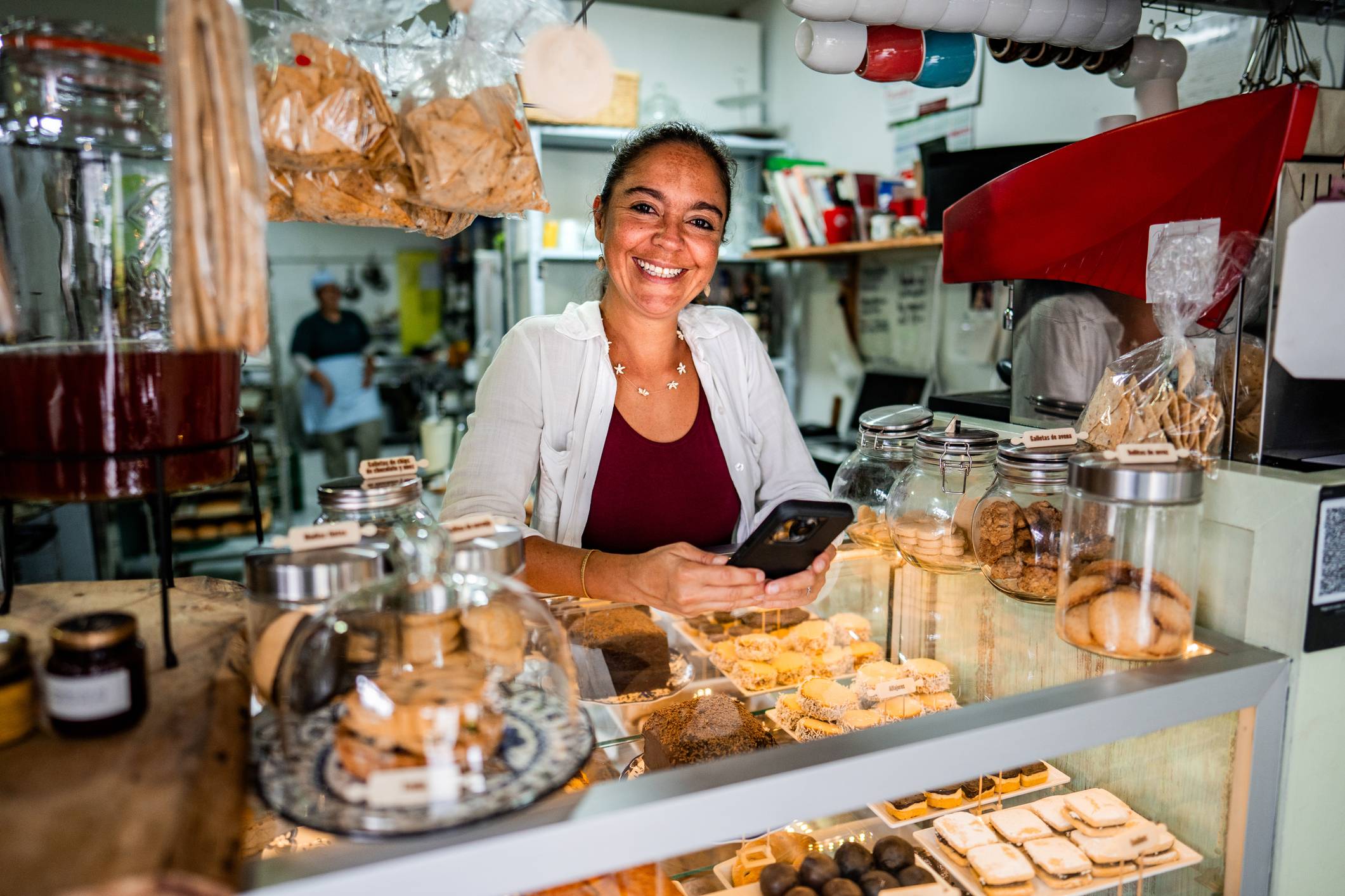 Mulher sorridente com celular atrás do balcão de uma padaria, rodeada por doces, biscoitos e bebidas.