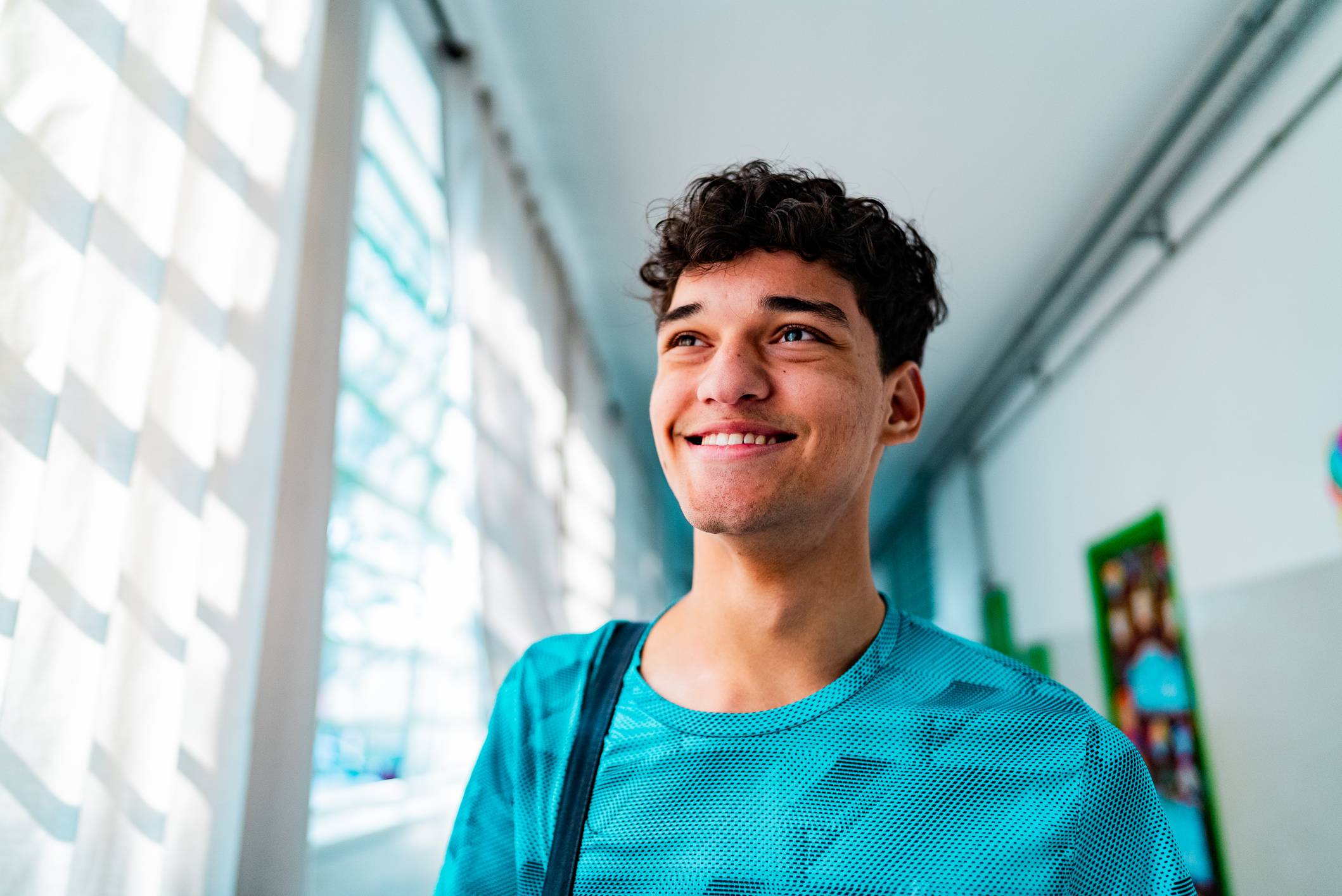 Jovem feliz com cabelo encaracolado sorrindo em corredor iluminado por janelas