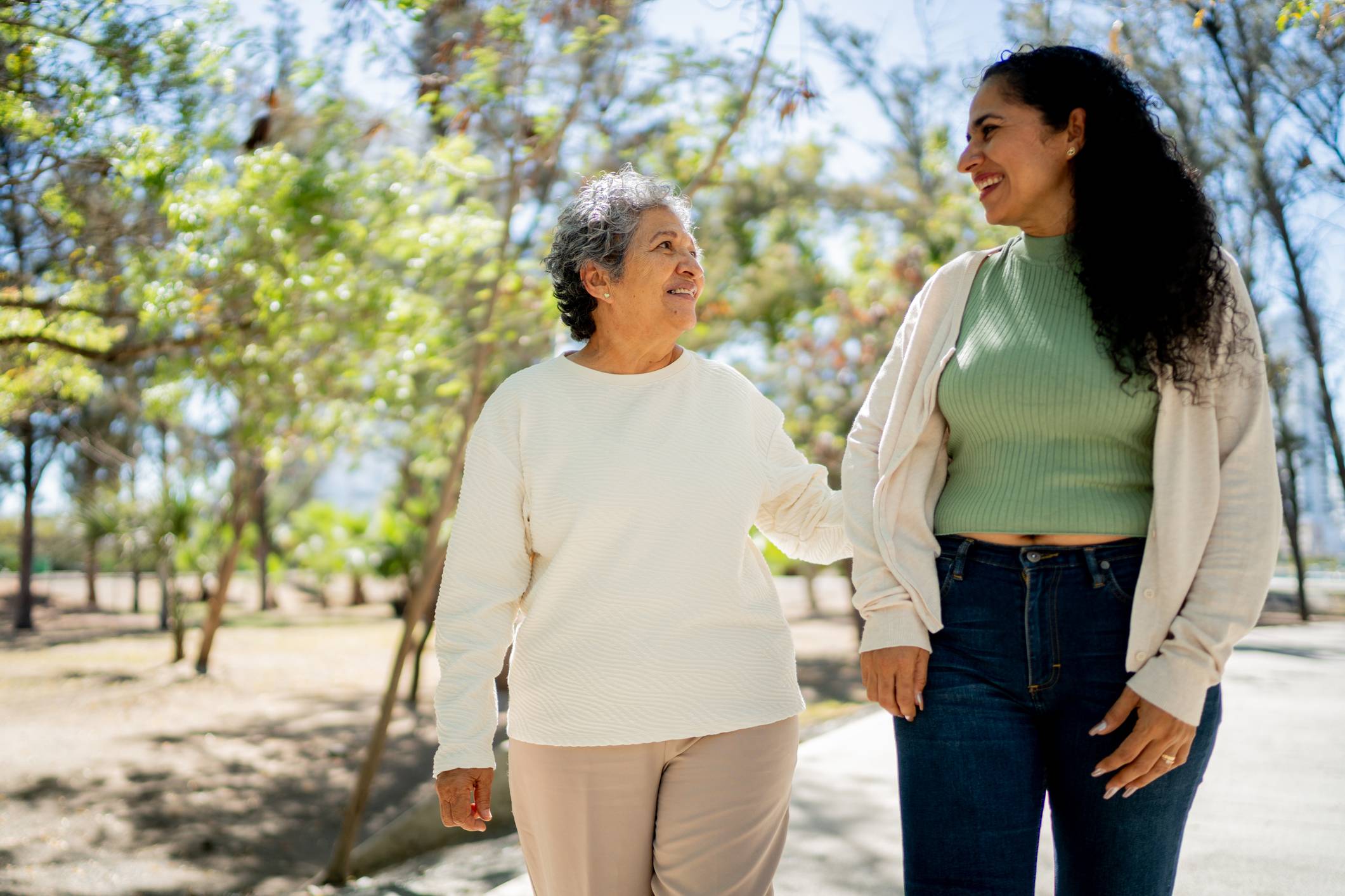 Duas mulheres sorrindo, uma idosa e uma jovem, caminhando de mãos dadas em um parque ensolarado.