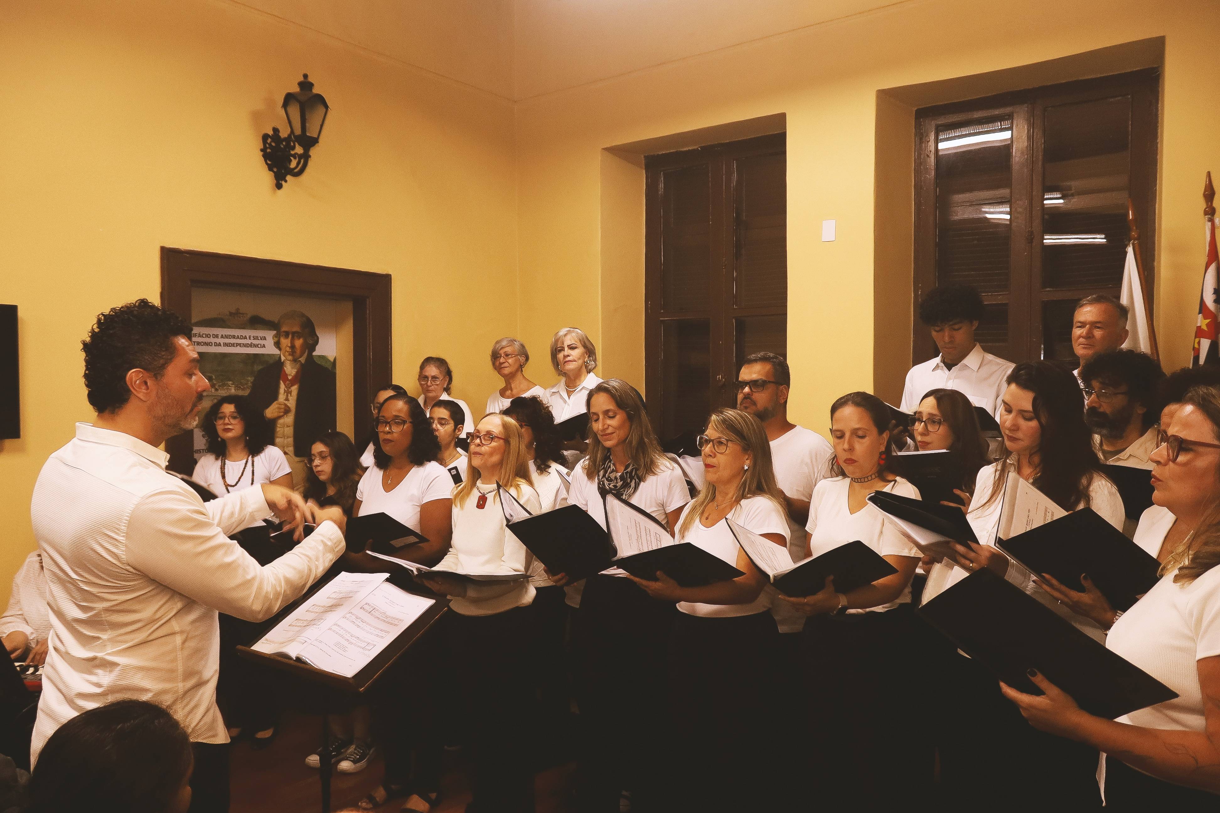 Maestro conduz coral diverso cantando com partituras. Ao fundo, quadro histórico de Bonifácio de Andrada e Silva.