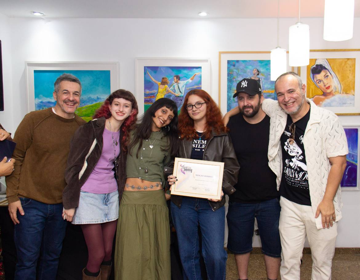 Seis pessoas sorridentes, incluindo uma jovem com certificado, posam em galeria de arte com pinturas coloridas ao fundo.
