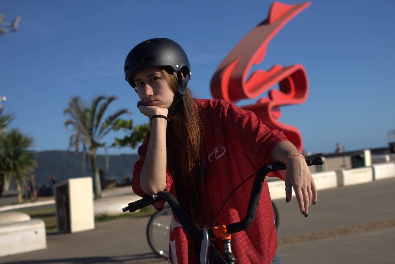 Jovem de capacete preto e camisa xadrez vermelha, apoiada em bicicleta com escultura vermelha e palmeiras ao fundo.