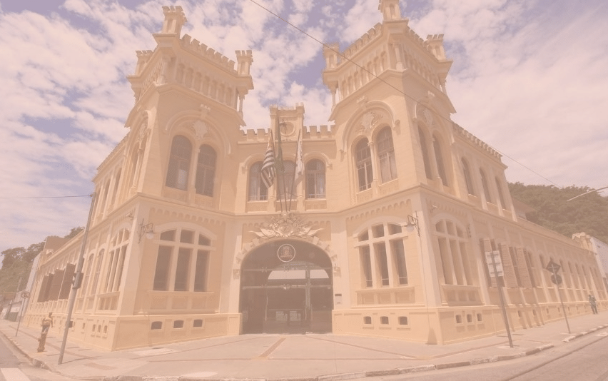 Prédio histórico amarelo da Câmara Municipal de Santos com duas torres ameias e bandeiras acima da entrada arqueada, sob céu azul com nuvens.