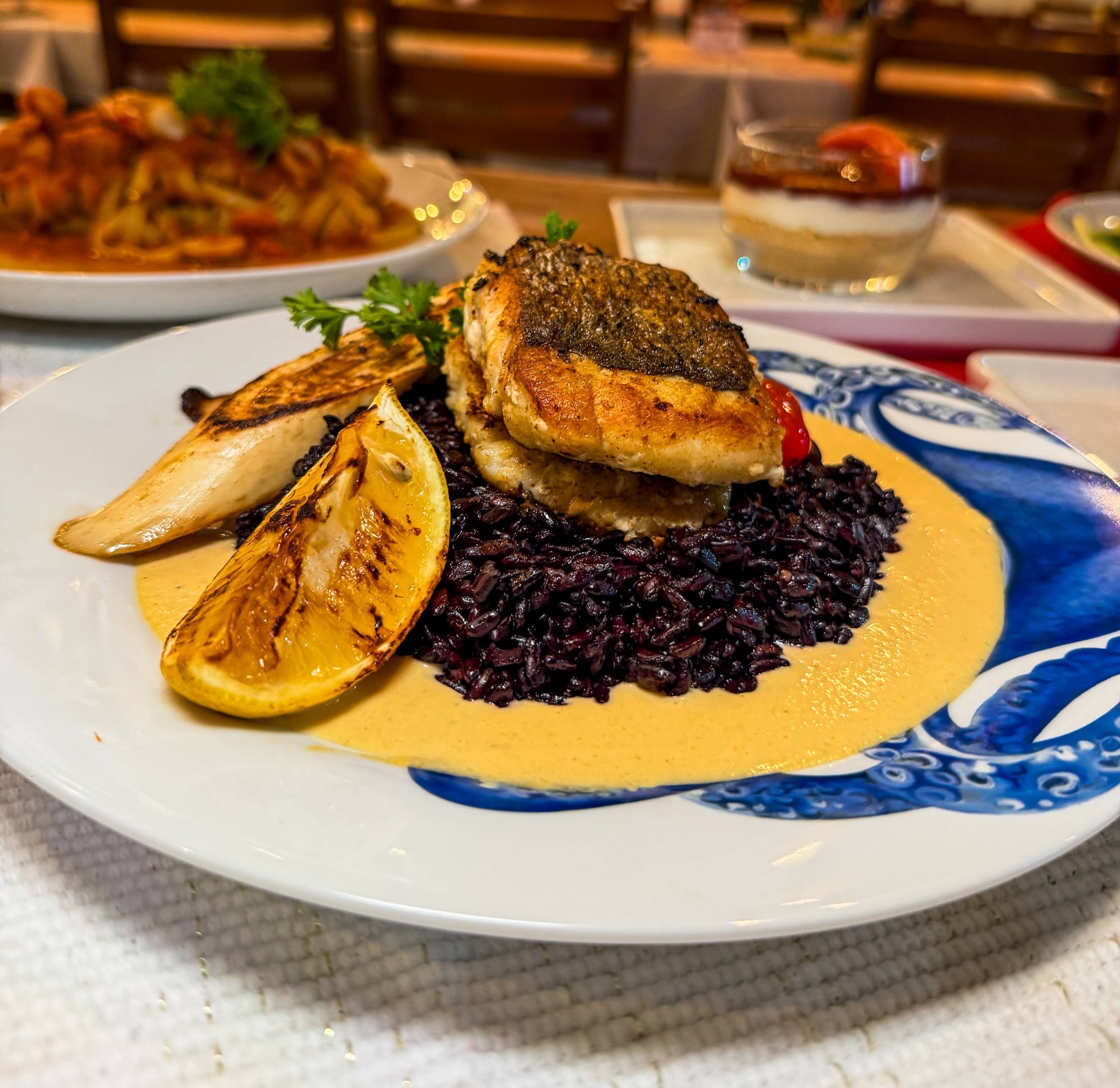 Peixe grelhado com pele crocante servido sobre arroz preto e molho cremoso, cogumelos, limão grelhado