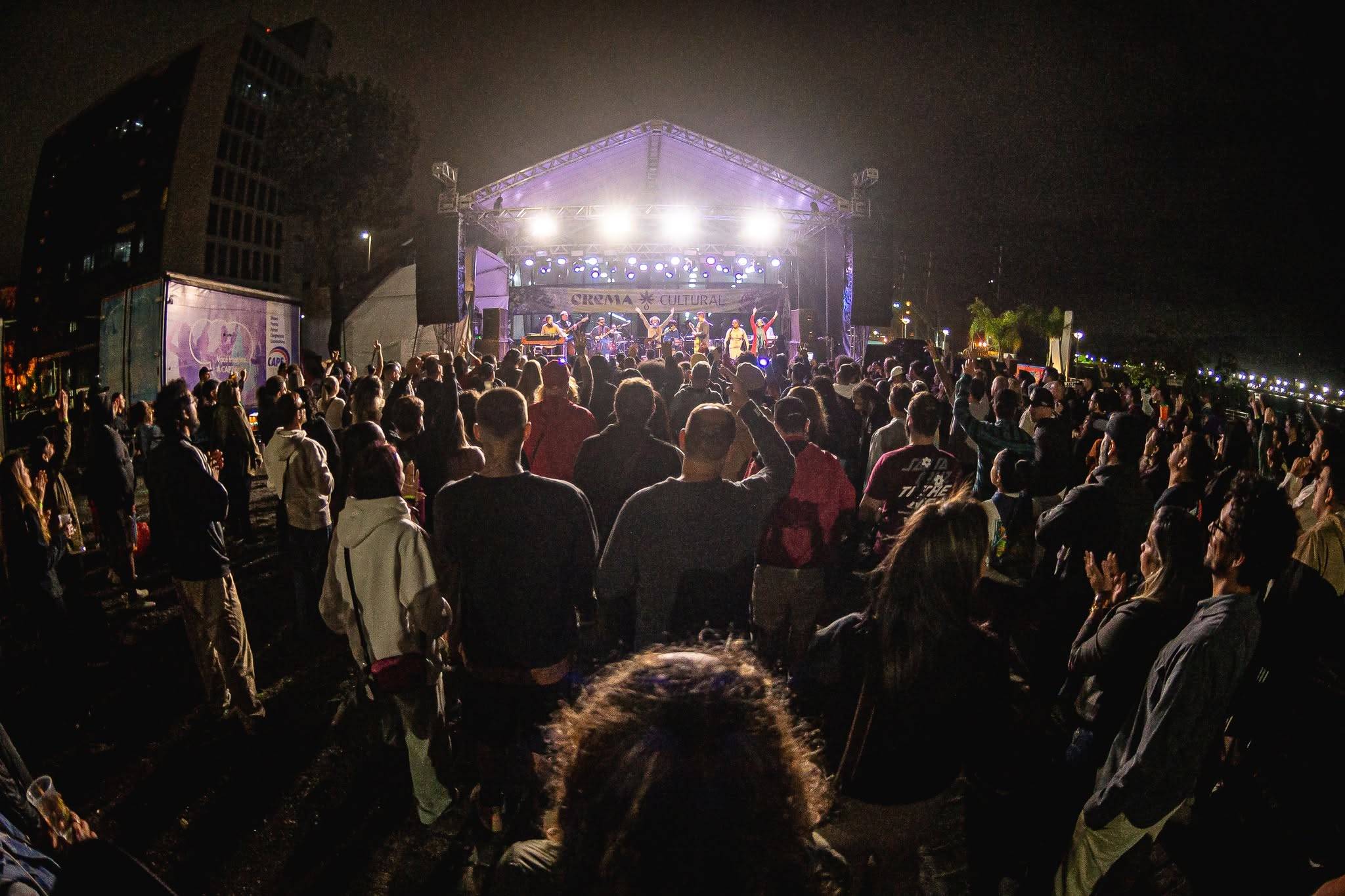 Multidão em um show de música ao vivo noturno, de costas para a câmera, olhando para um palco iluminado com uma banda se apresentando. Um prédio e um caminhão estão à esquerda.