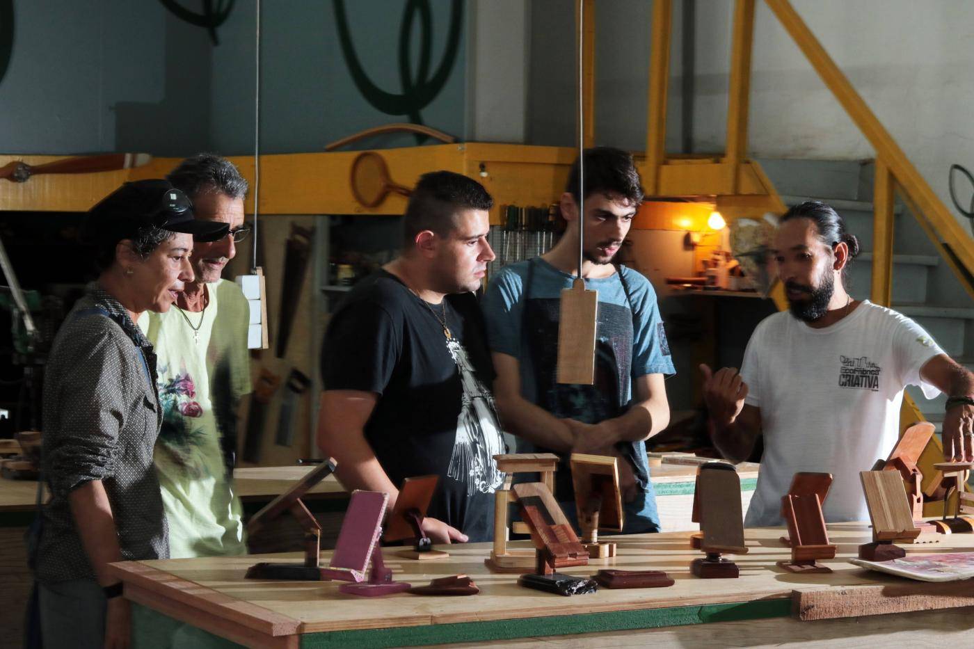 Grupo de pessoas em oficina de marcenaria, observando e discutindo diversos modelos de suportes de celular em madeira artesanais sobre uma mesa.
