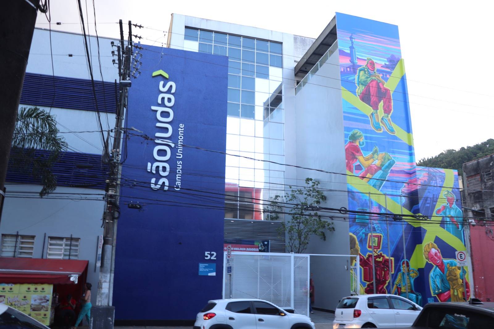 Fachada azul do campus São Judas Unimonte com seu nome em branco e um grande mural colorido na lateral. Carros e poste com fios visíveis na frente.