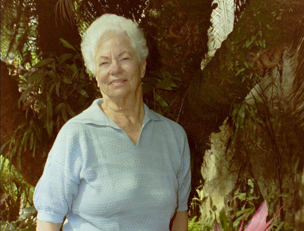 Mulher idosa sorrindo com cabelo branco e camisa polo azul-claro, em meio a folhagem verde tropical