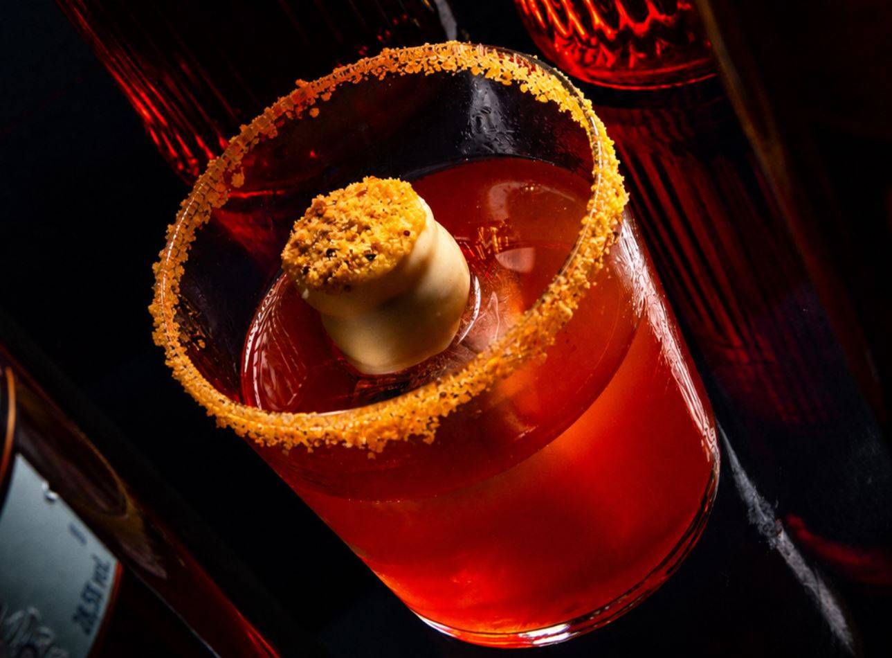 Coquetel vermelho vibrante em copo baixo com borda crocante e garnish esférico cremoso com crumble dourado.