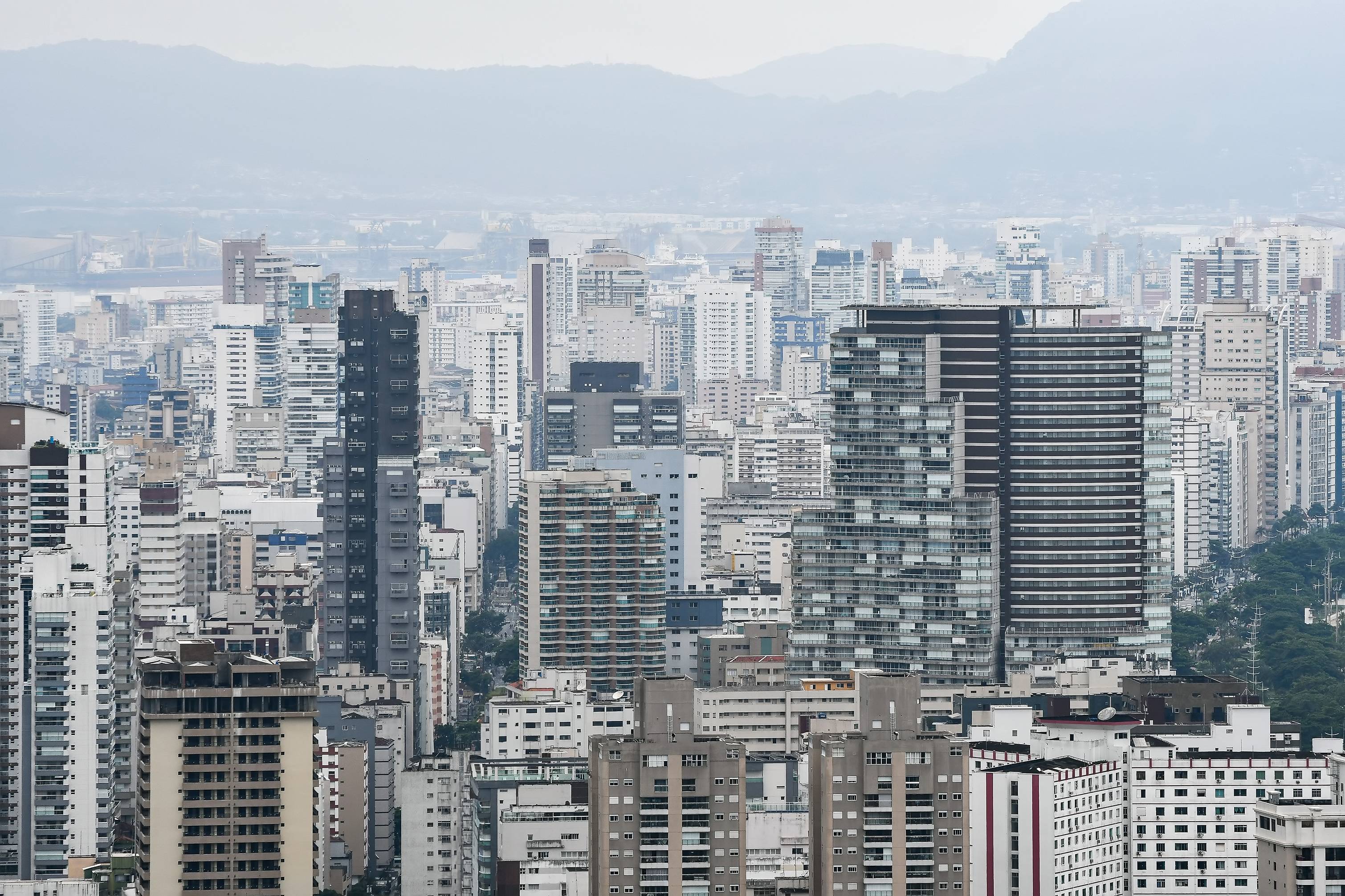 Panorama urbano denso com arranha-céus modernos, revelando montanhas e estruturas portuárias distantes em dia nublado.