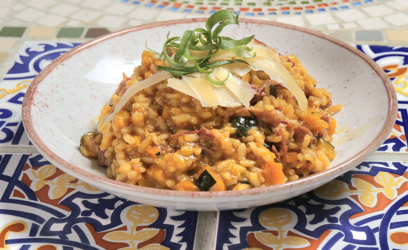 Risoto cremoso de abóbora com carne desfiada e queijo parmesão ralado, guarnecido com ervas frescas em tigela decorada.