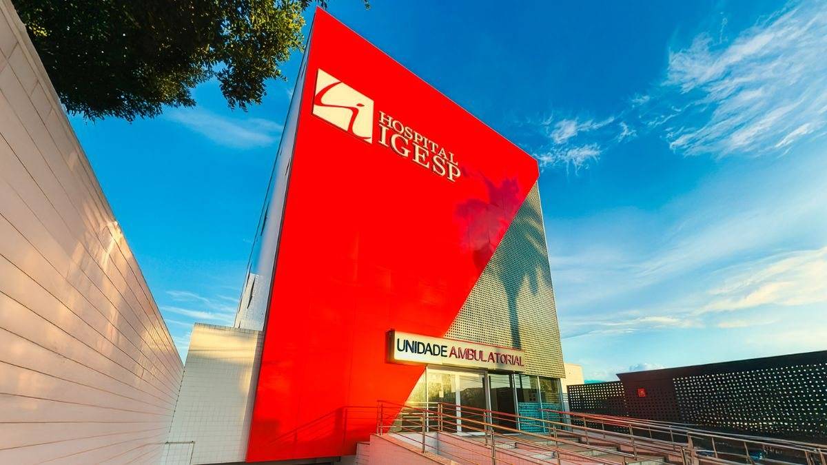 Fachada moderna e angular do Hospital IGESP em vermelho vibrante com a sinalização 'Unidade Ambulatorial', sob um céu azul claro.
