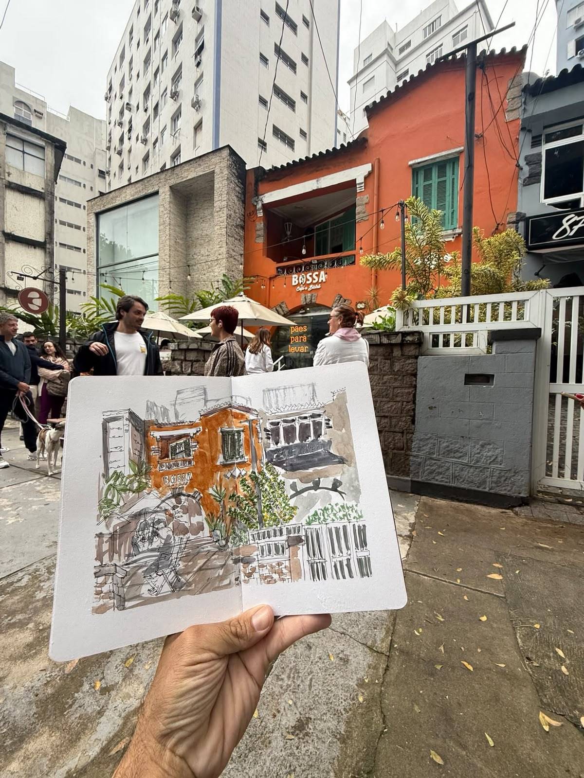 Mão segurando sketchbook com desenho de rua aquarelado e a cena real com prédio laranja do Bossa Café ao fundo.